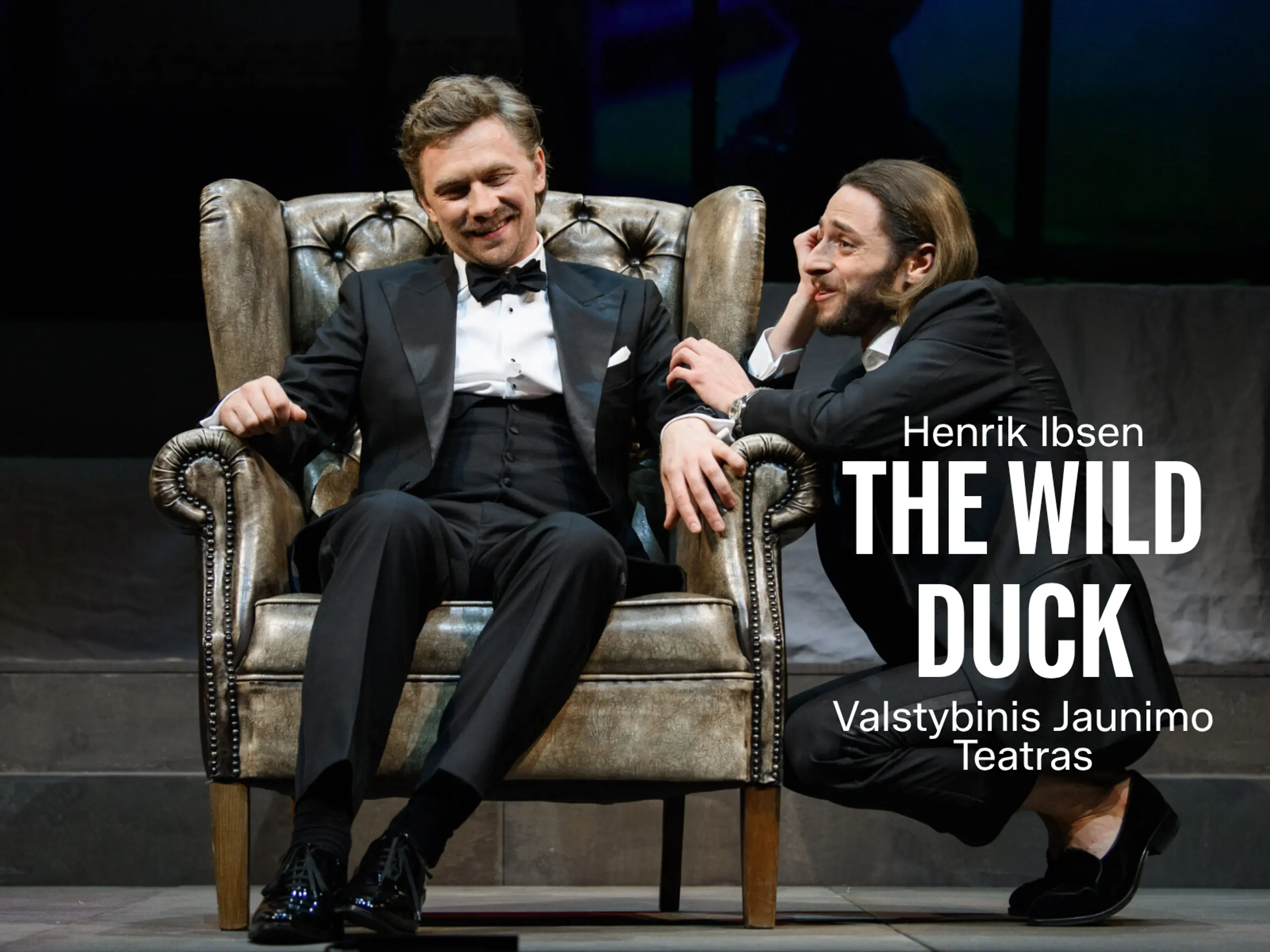 Smilende mann i smoking sitter i en skinnlenestol mens en annen mann på huk lener seg mot armlenet under en samtale. Med tittel "Henrik Ibsen The Wild Duck"