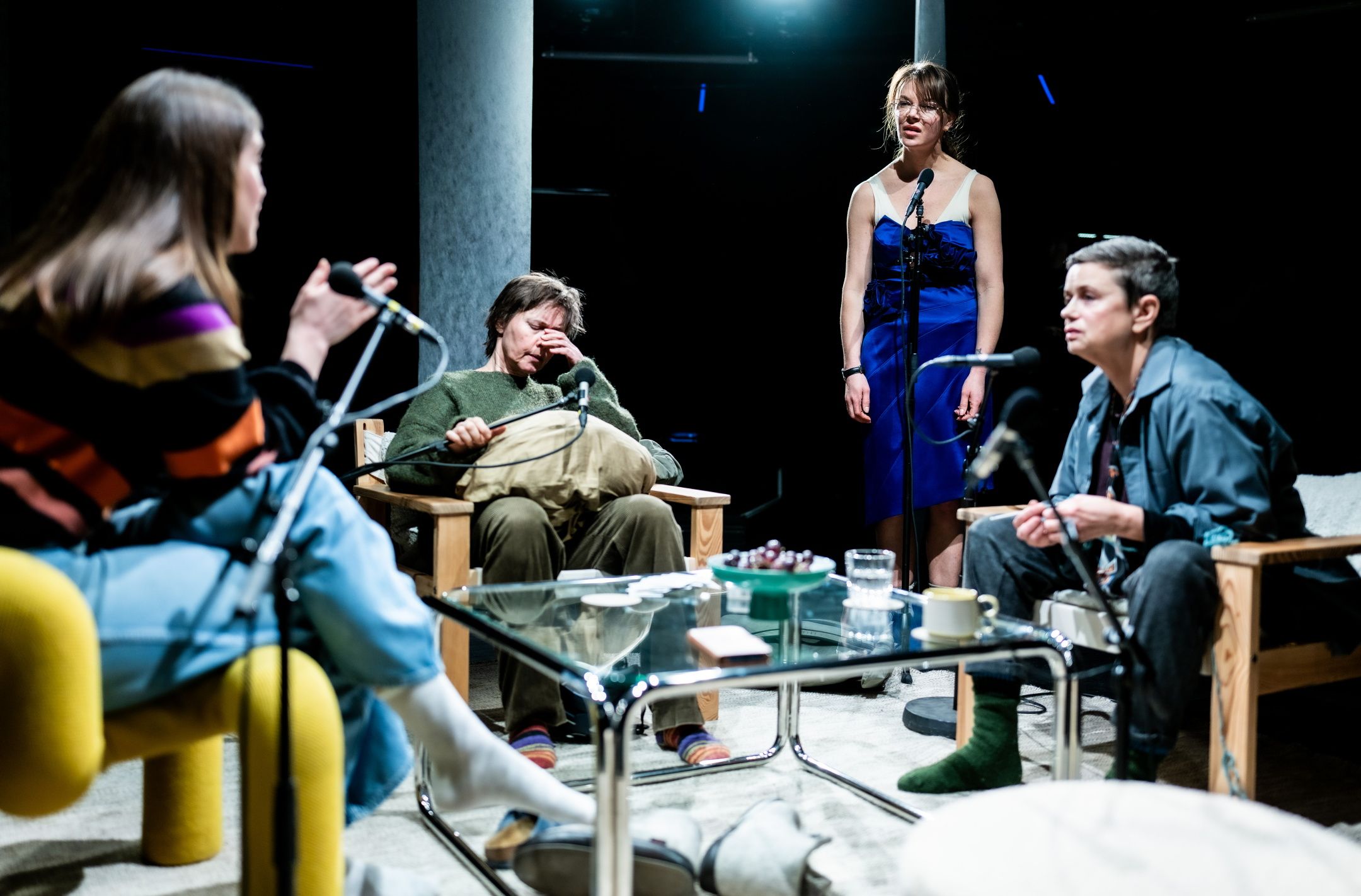 Fire personer rundt et bord i scenisk setting, én står i blå kjole, én gnir seg i øyet.