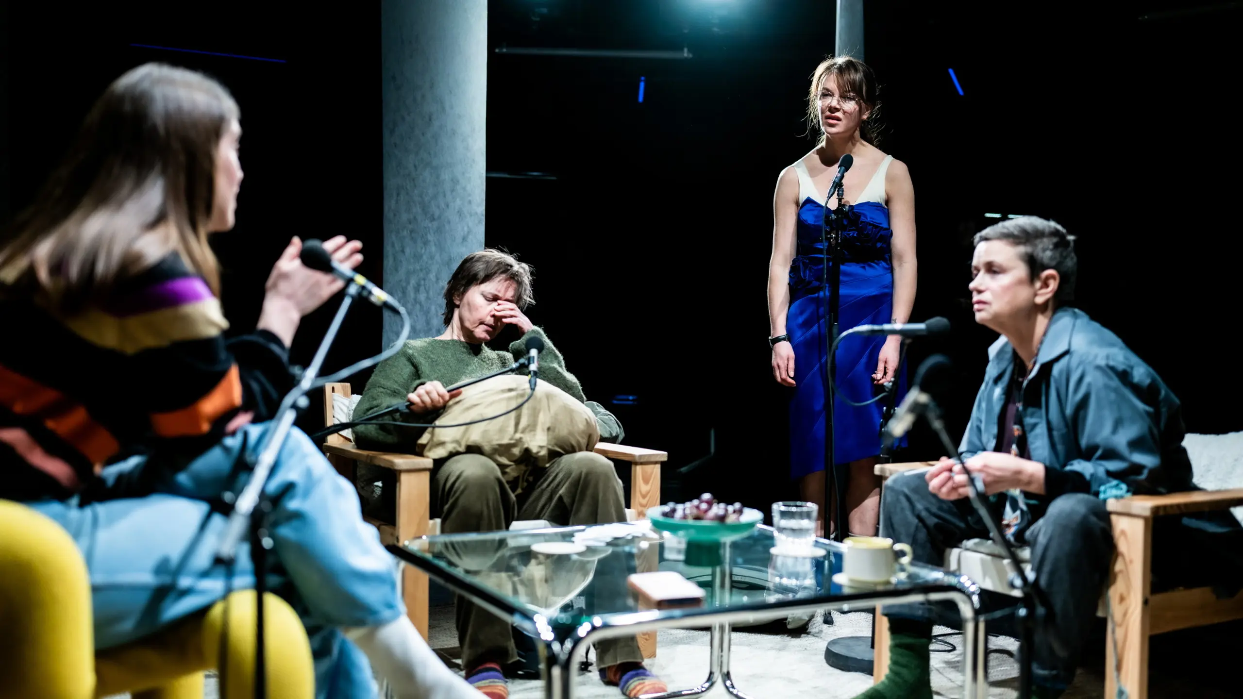 Fire personer rundt et bord i scenisk setting, én står i blå kjole, én gnir seg i øyet.