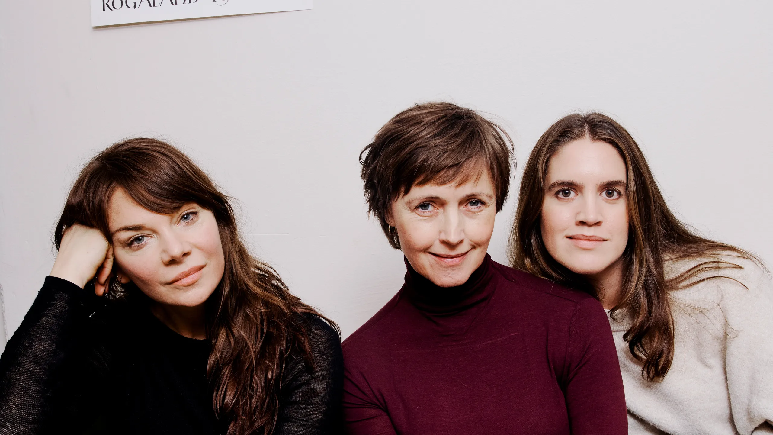 Tre kvinner sitter i en grønn sofa mot en hvit vegg.