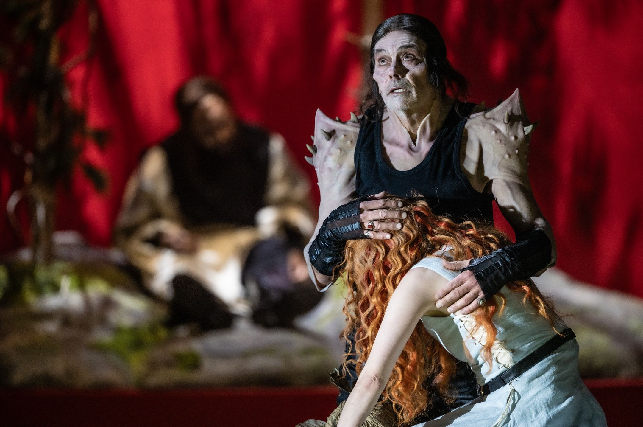 En mann i svart drakt og piggete skuldre holder beskyttende rundt en kvinne med rødt hår som faller mot ham.