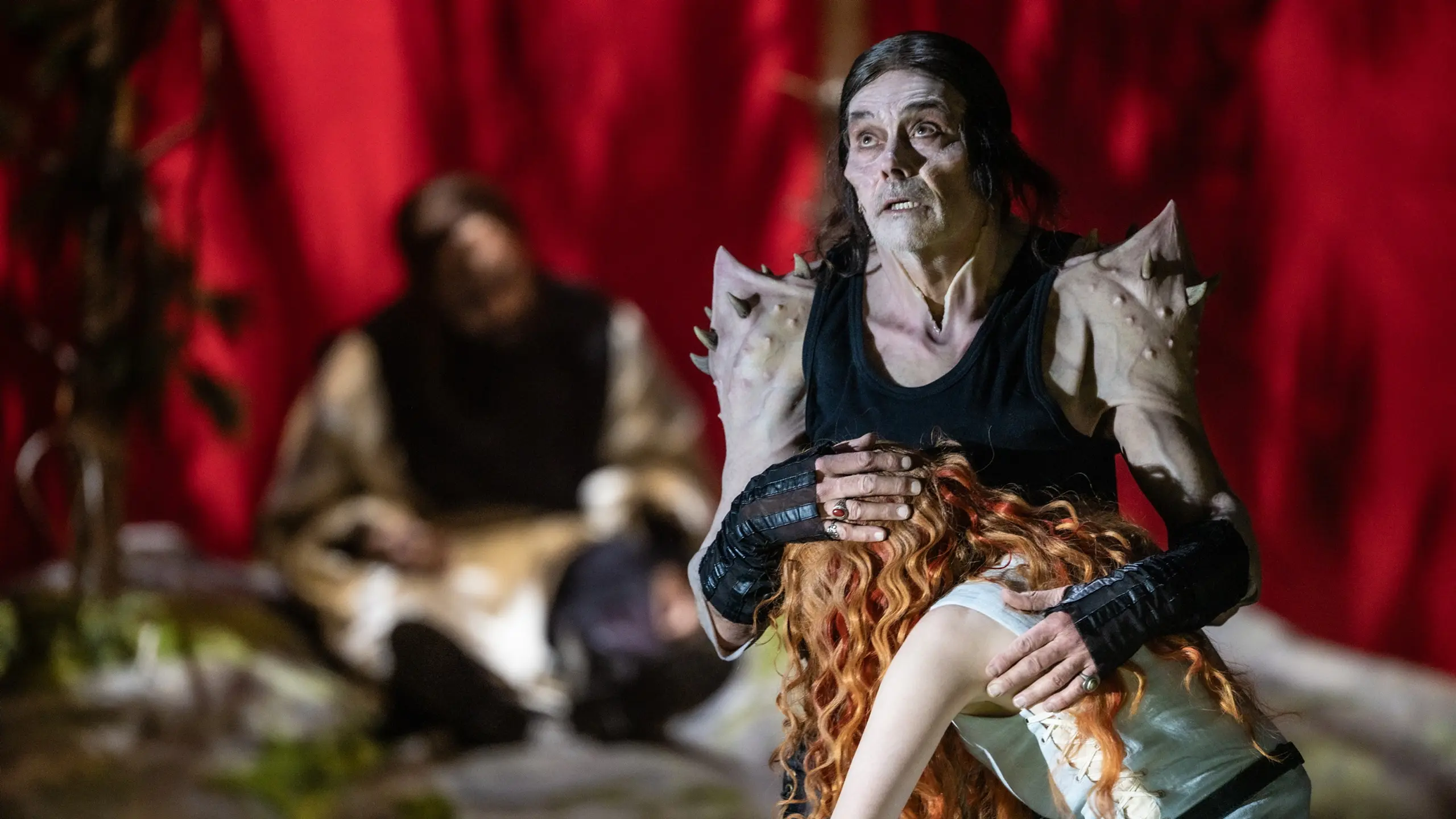 En mann i svart drakt og piggete skuldre holder beskyttende rundt en kvinne med rødt hår som faller mot ham.