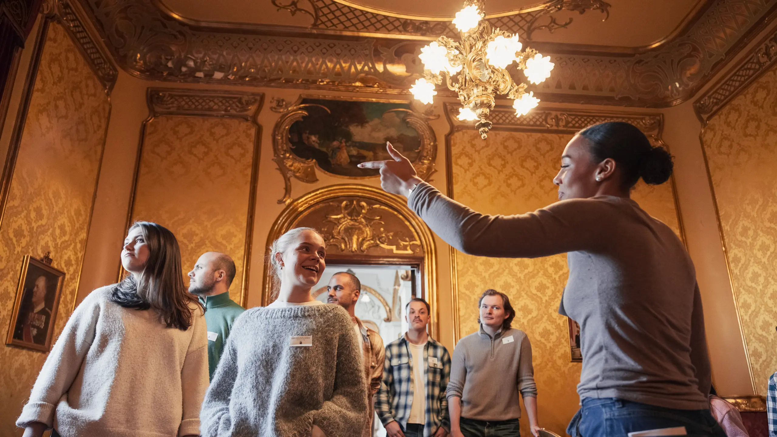 Omviser gestikulerer og snakker til en gruppe voksne i et rom med gyldne vegger og lysekrone.