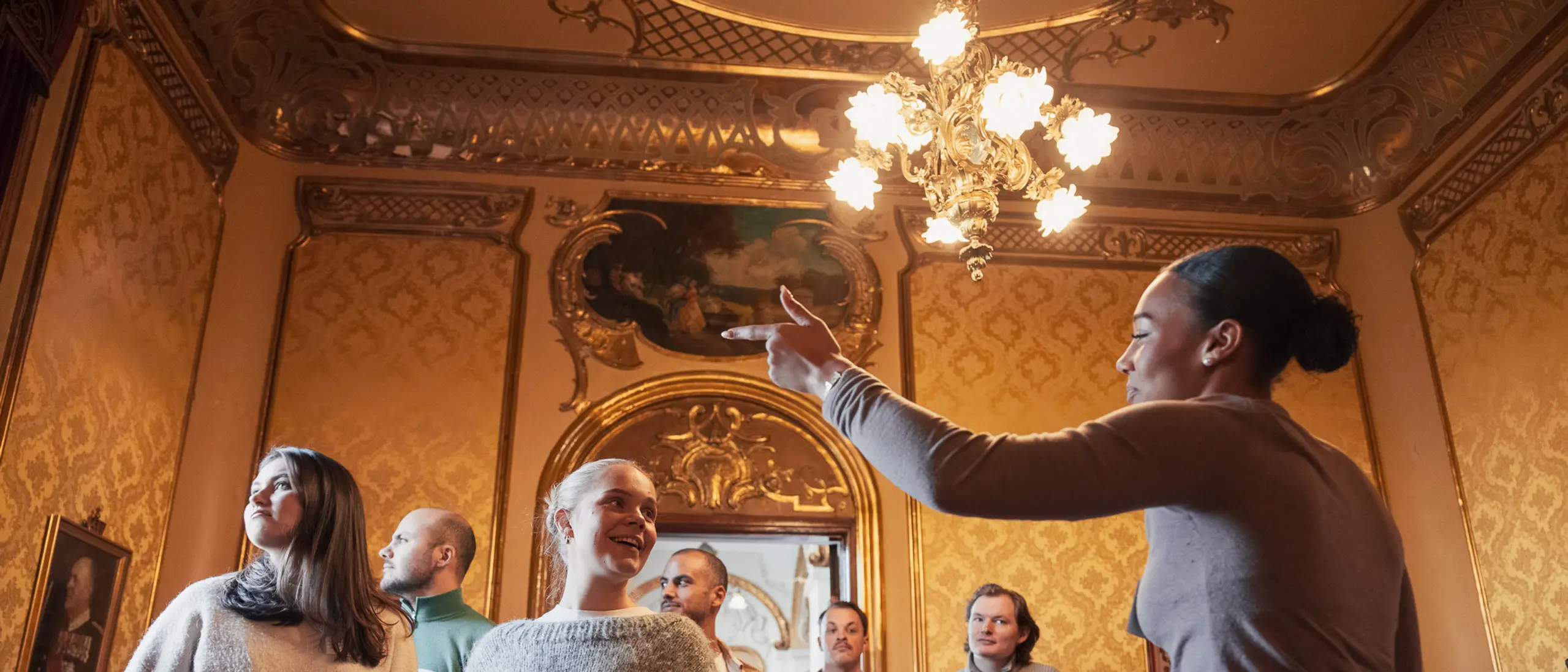Omviser gestikulerer og snakker til en gruppe voksne i et rom med gyldne vegger og lysekrone.