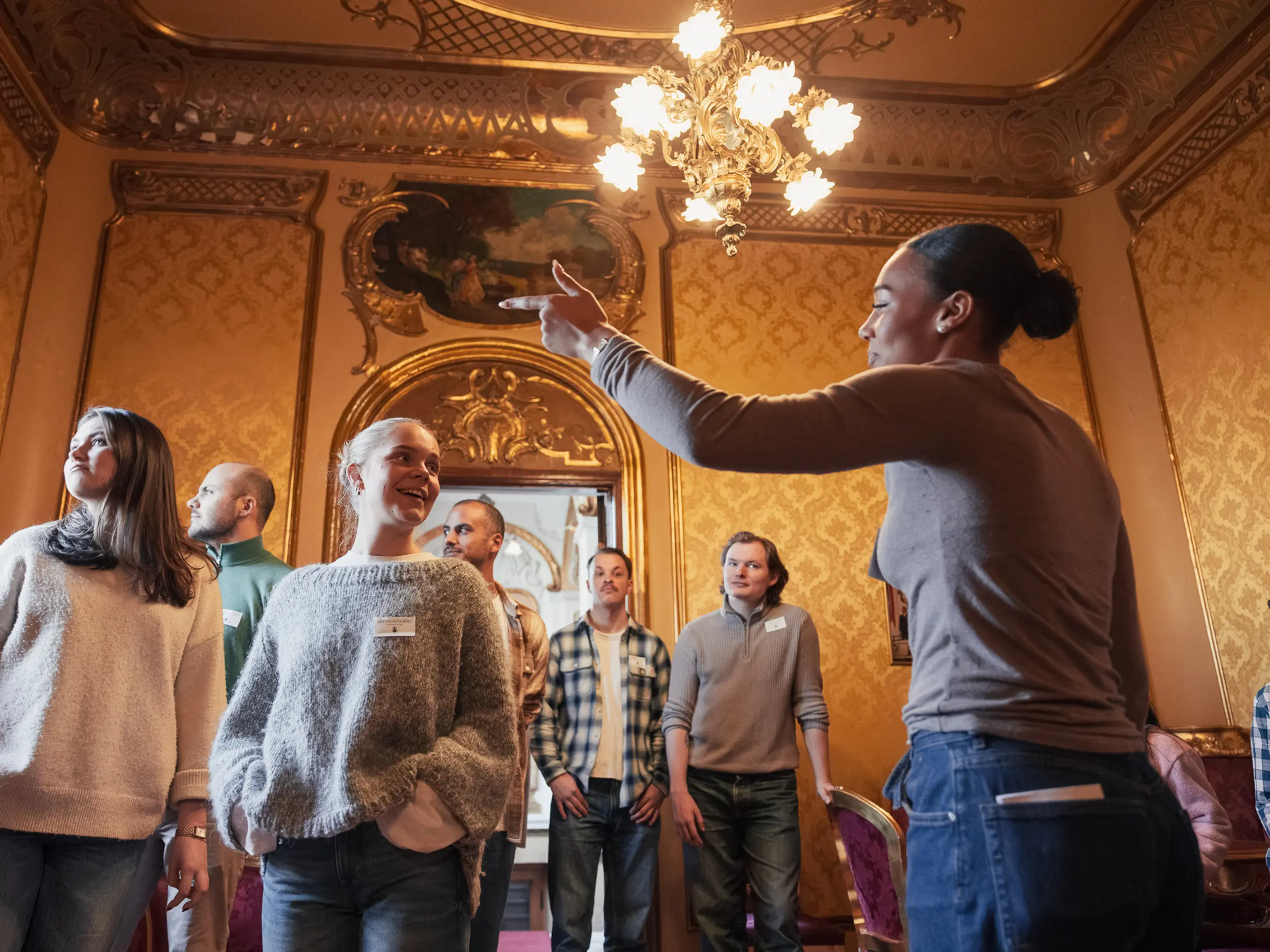 Omviser gestikulerer og snakker til en gruppe voksne i et rom med gyldne vegger og lysekrone.