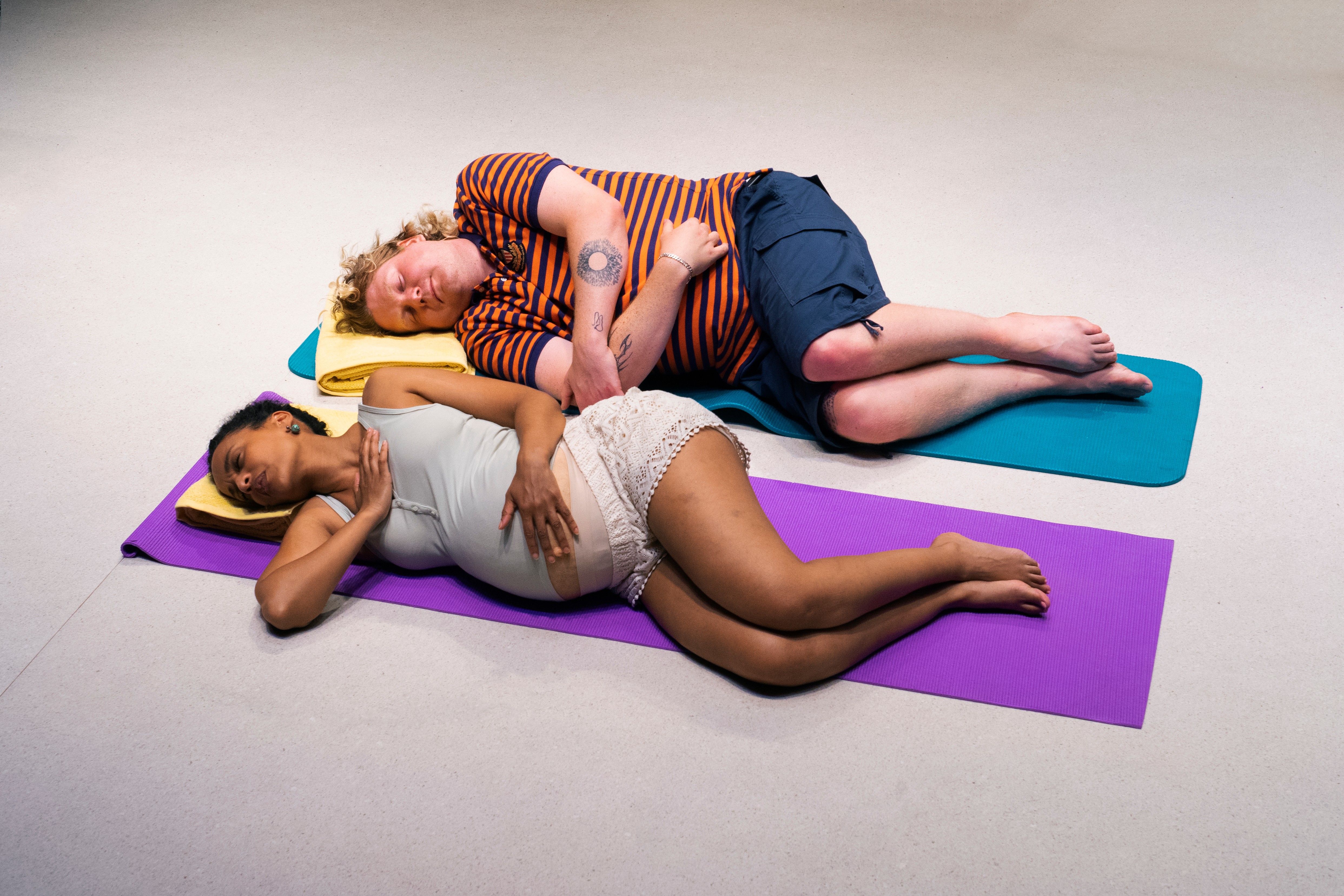 Et gravid par ligger på yogamatter, kvinnen på lilla matte og mannen på blå, begge med øynene lukket.