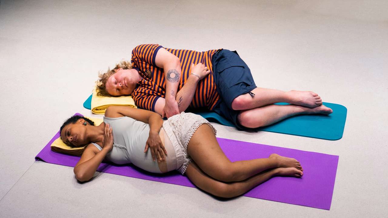 Et gravid par ligger på yogamatter, kvinnen på lilla matte og mannen på blå, begge med øynene lukket.