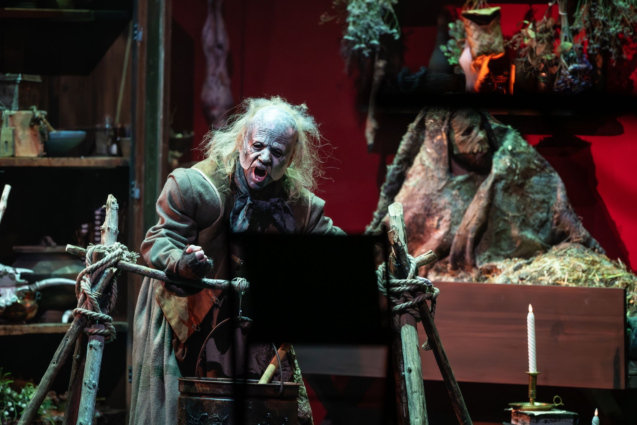 En skuespiller med vill parykk og grotesk sminke roper ut mot publikum i et mørkt og rotete heksekjøkken-scenografi.