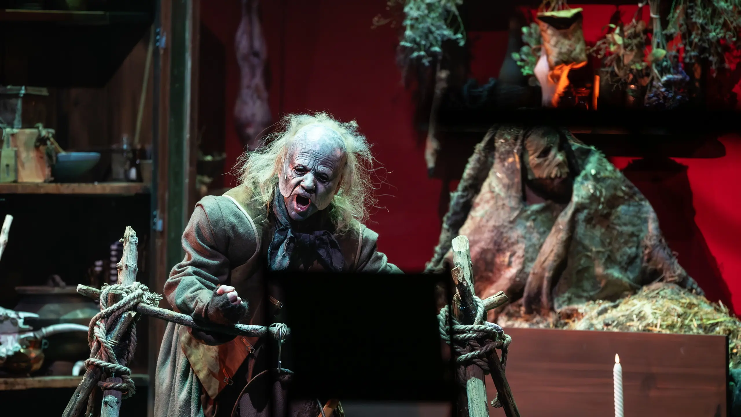 En skuespiller med vill parykk og grotesk sminke roper ut mot publikum i et mørkt og rotete heksekjøkken-scenografi.