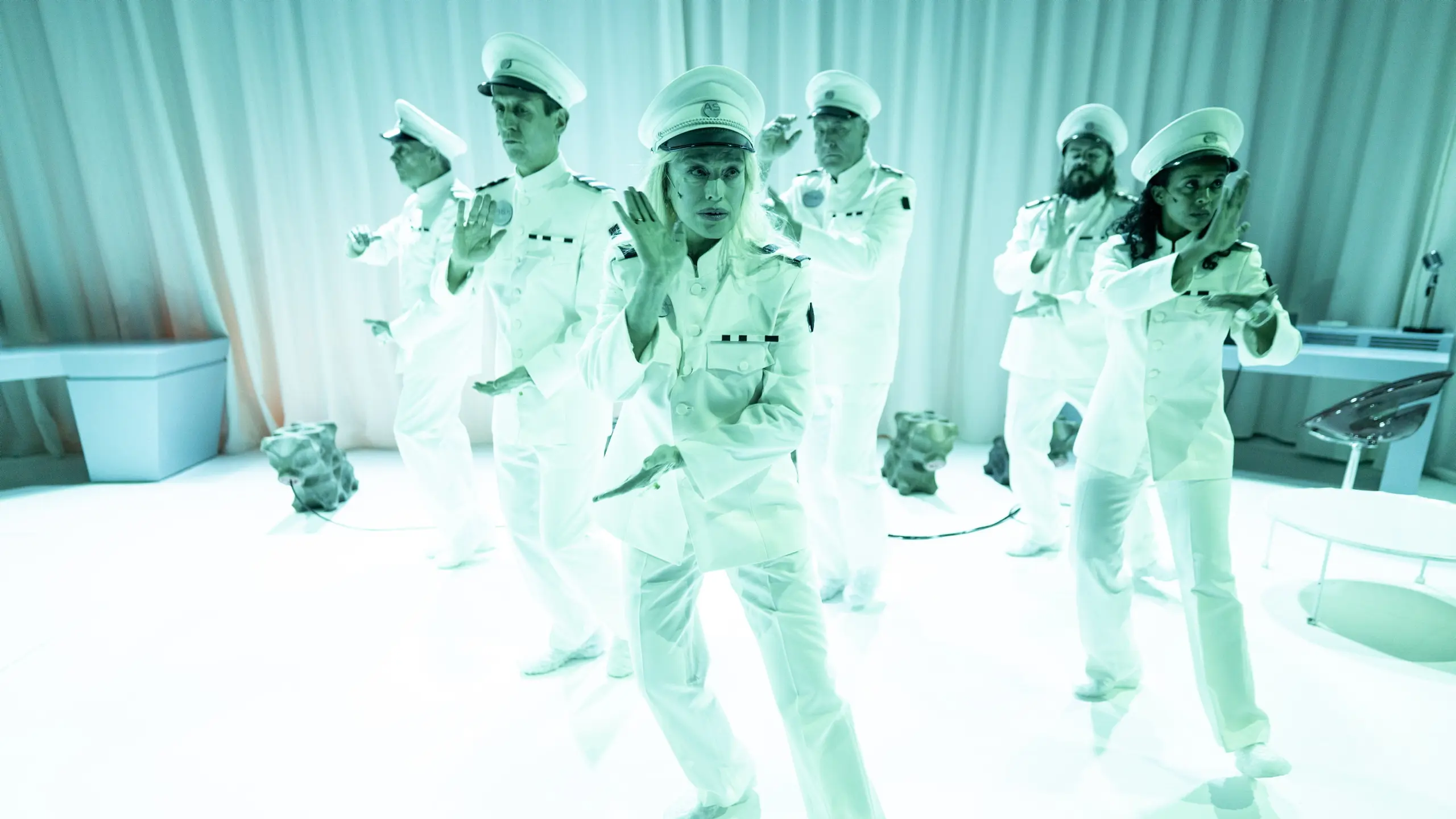Seks personer i hvite uniformer utfører synkroniserte håndbevegelser i et lyst, futuristisk rom