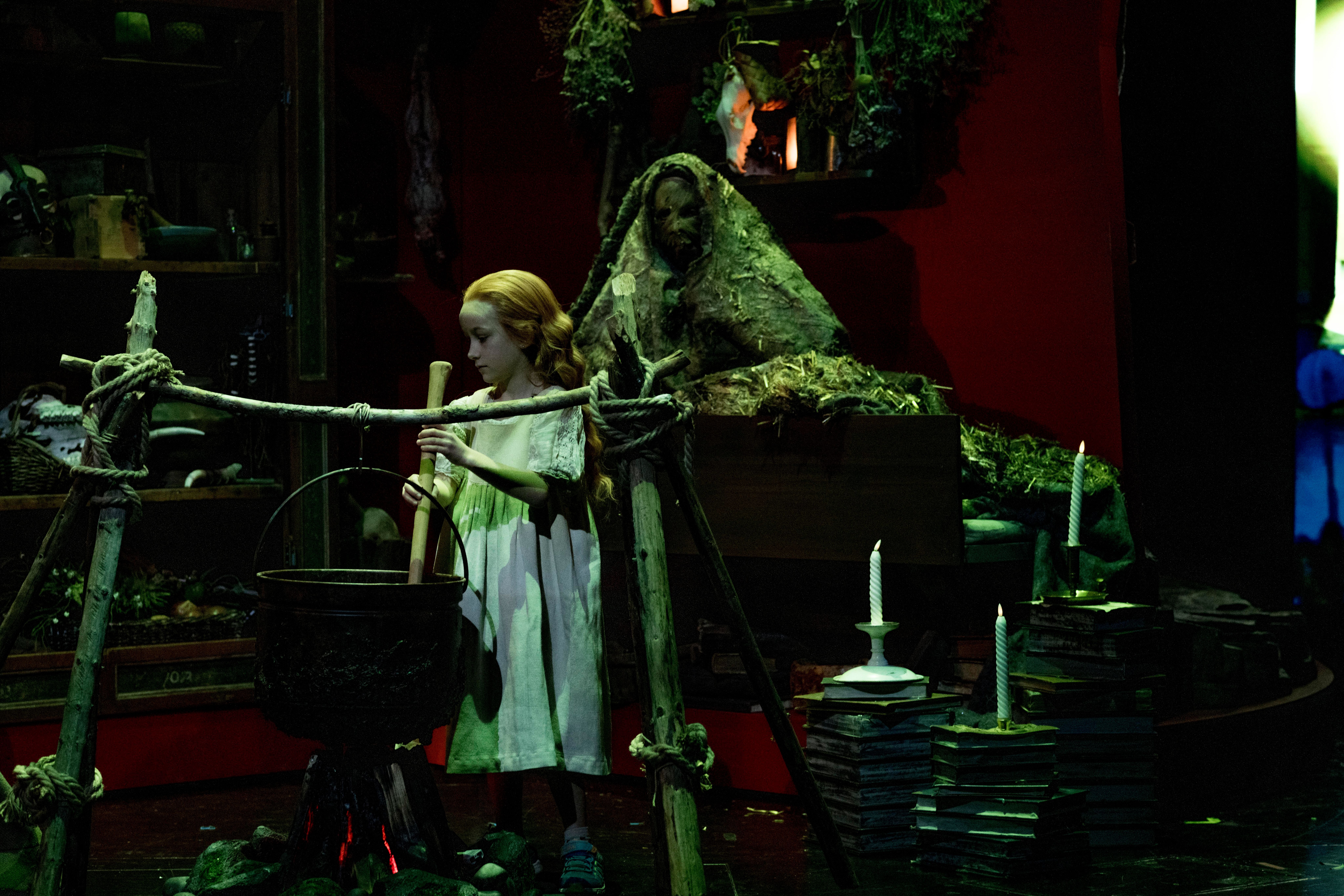 ung jente i hvit kjole som rører i en gryte over bål, omgitt av urter og en figur tildekket av mose i bakgrunnen.