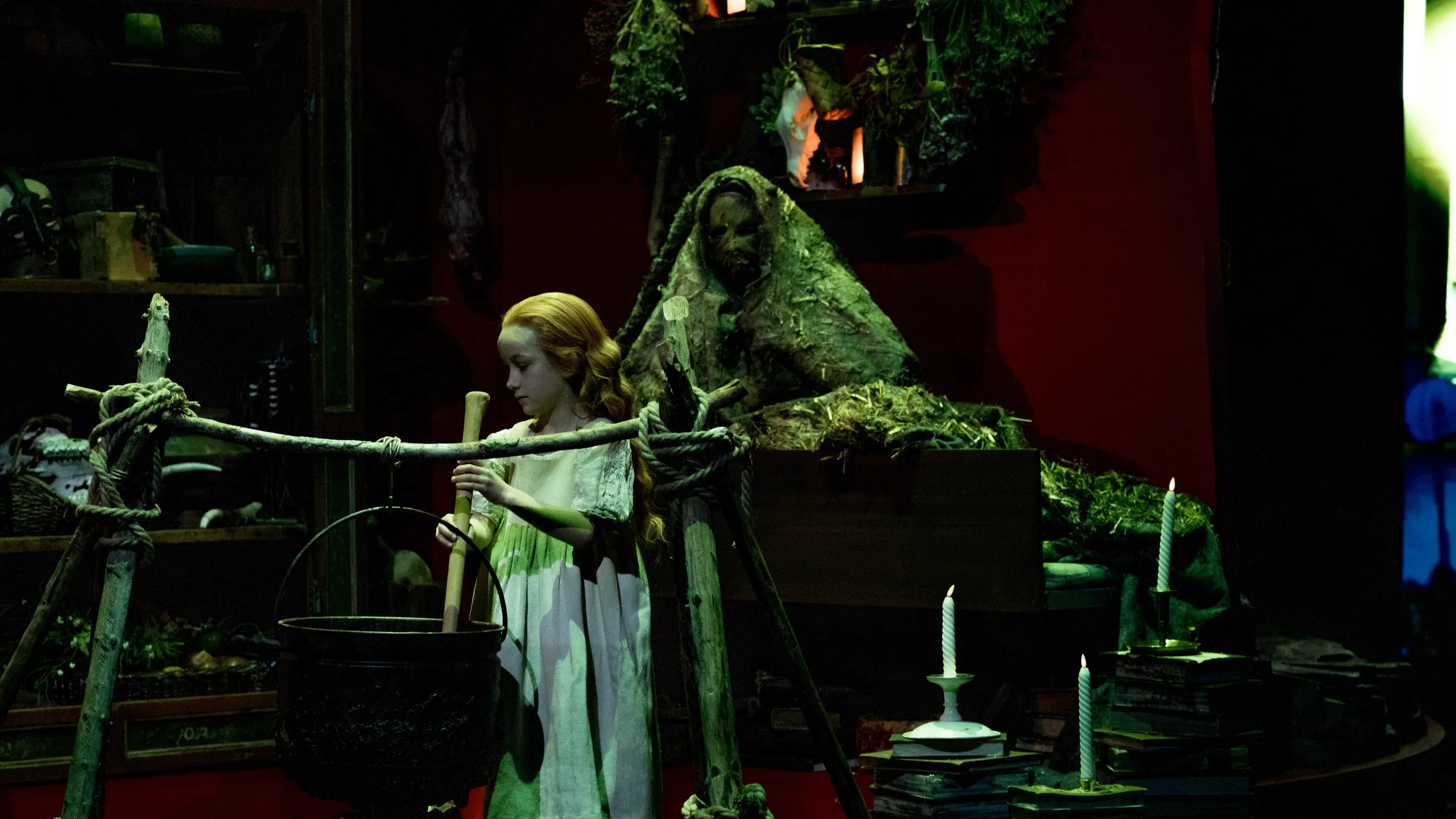 ung jente i hvit kjole som rører i en gryte over bål, omgitt av urter og en figur tildekket av mose i bakgrunnen.