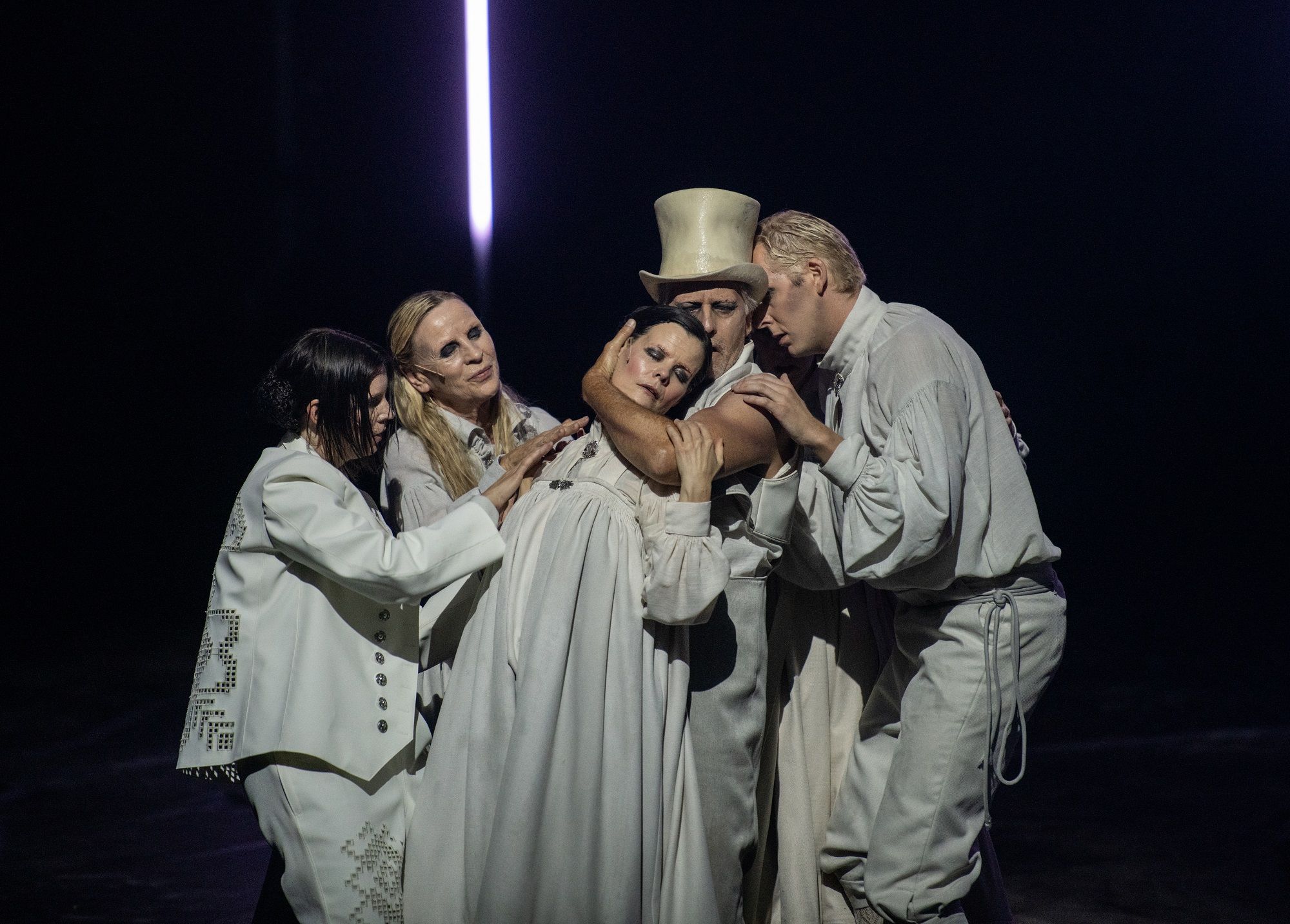 Gruppe på fem skuespillere i lyse kostymer, én med høy flosshatt, hvor en sentral figur holdes i armene på en mørklagt scene.