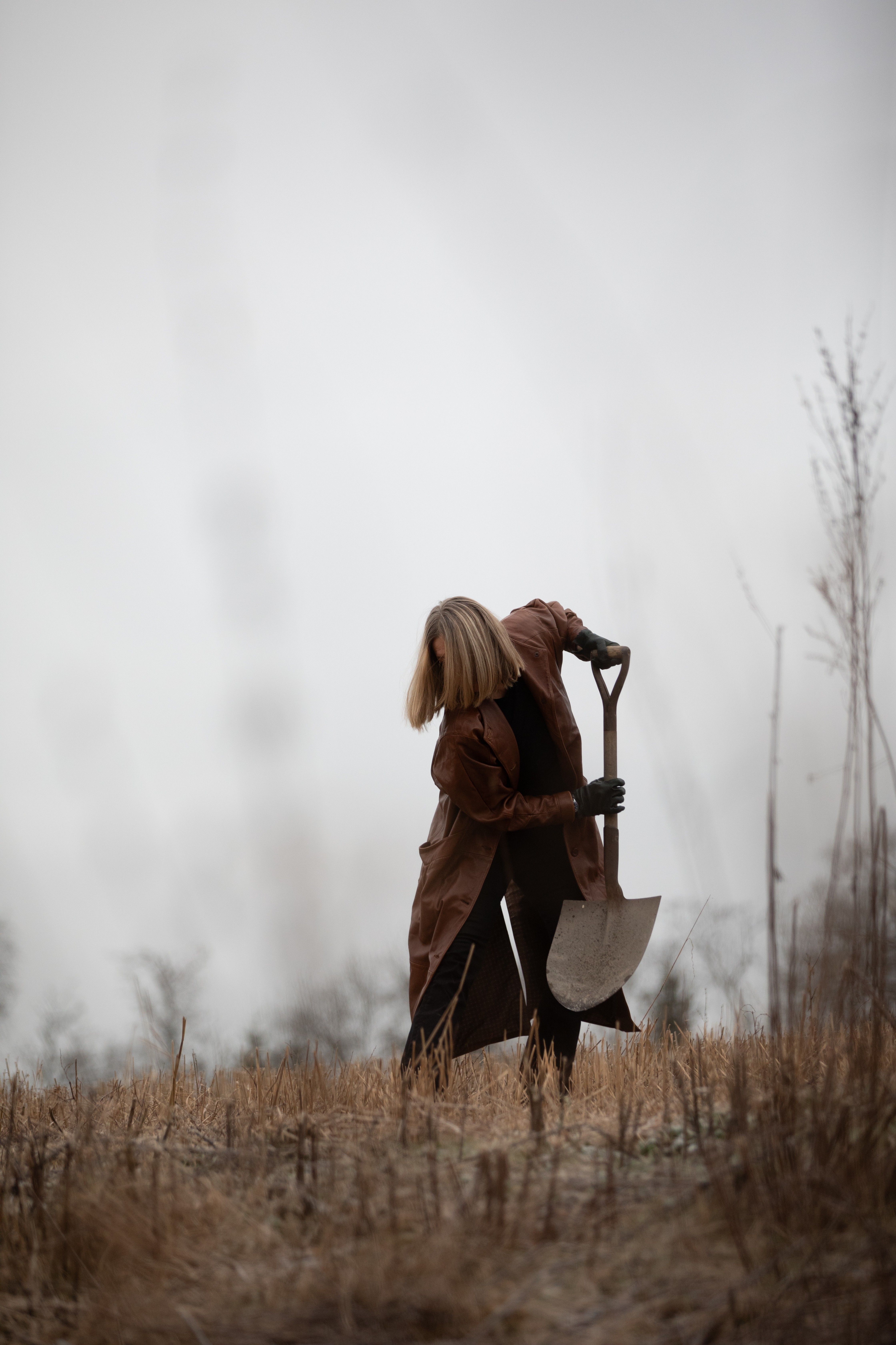 Person med blondt hår i brun frakk graver i en åker med en spade, mot en disig bakgrunn.