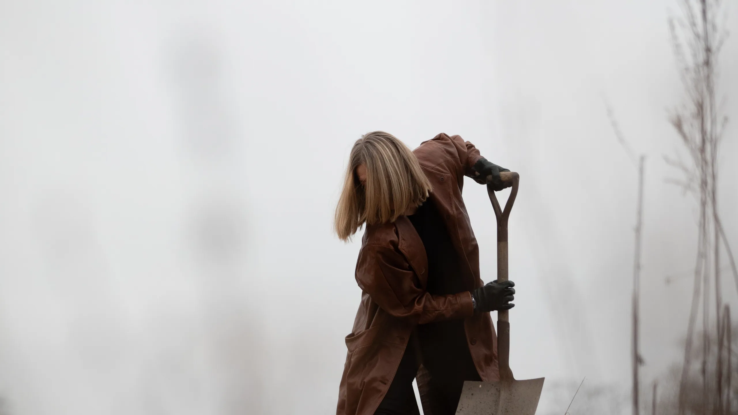 Person med blondt hår i brun frakk graver i en åker med en spade, mot en disig bakgrunn.