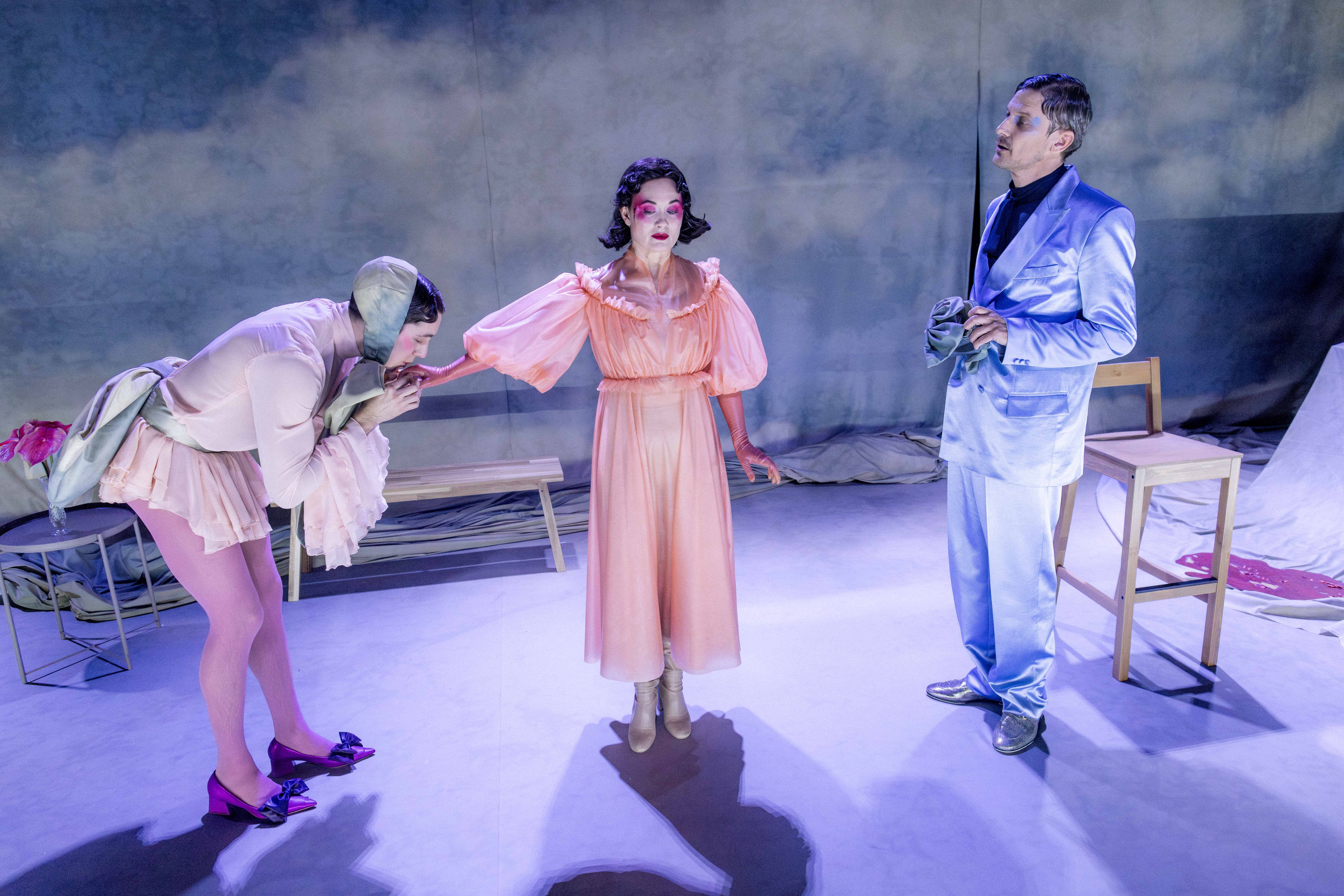 Tre personer på scenegulv; en i ferskenfarget kjole står mellom to personer, hvor en bøyer seg og kysser hånden hennes