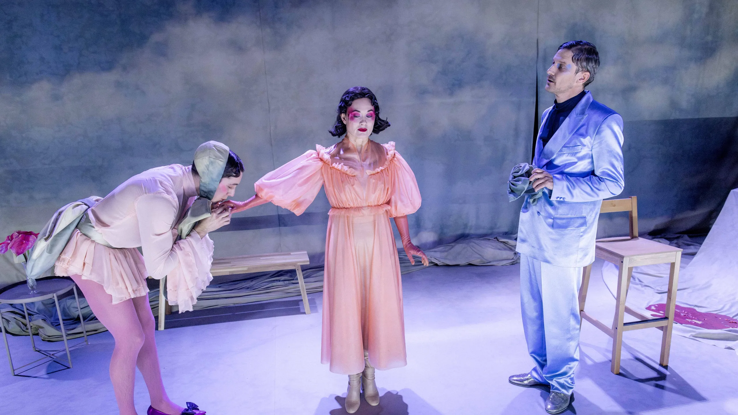 Tre personer på scenegulv; en i ferskenfarget kjole står mellom to personer, hvor en bøyer seg og kysser hånden hennes