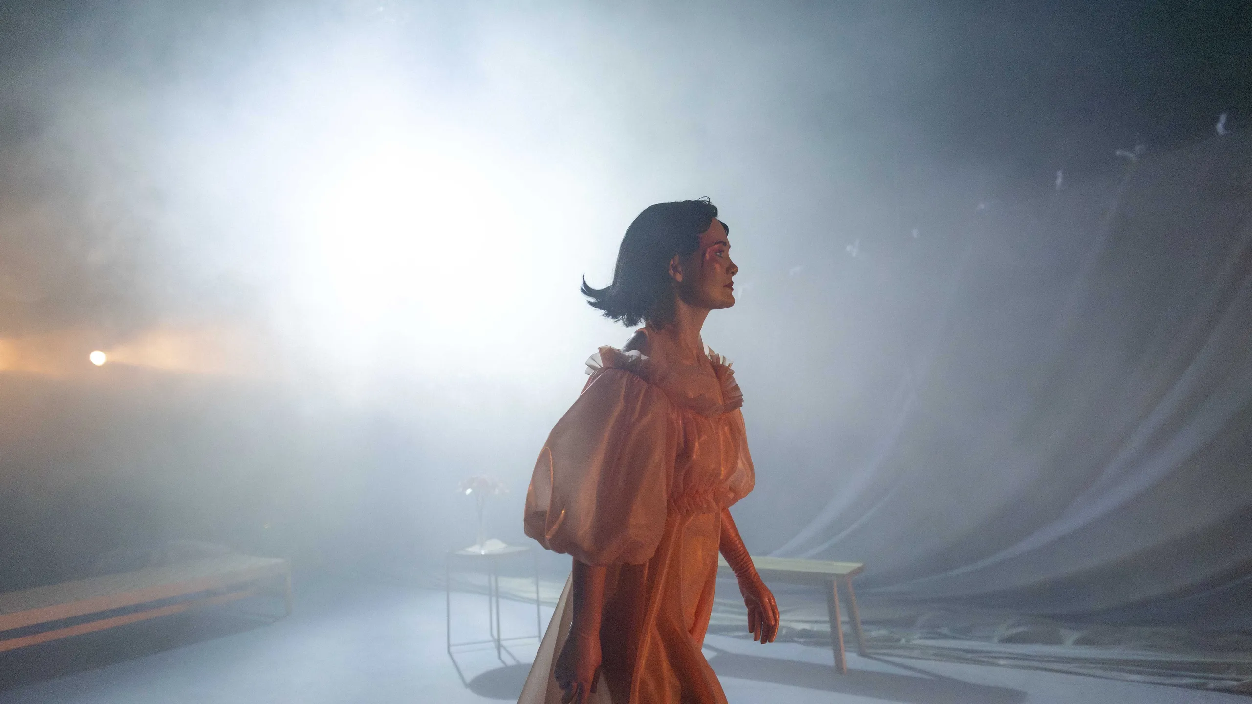 Person i ferskenfarget kjole går gjennom røykfylt scene med sterkt baklys