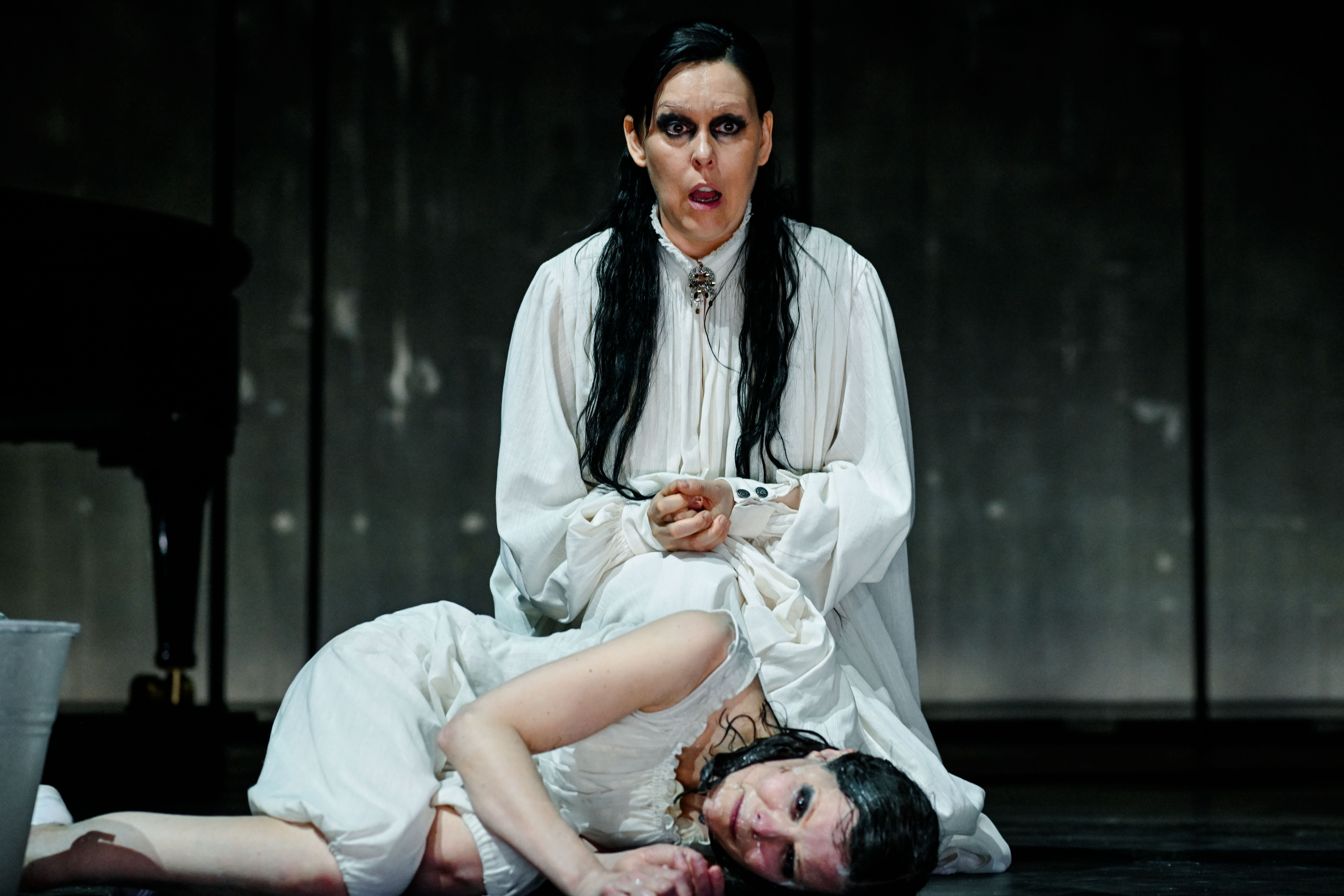 En person sitter på kne og synger bak en som ligger på gulvet og gråter, tilgriset i væske.