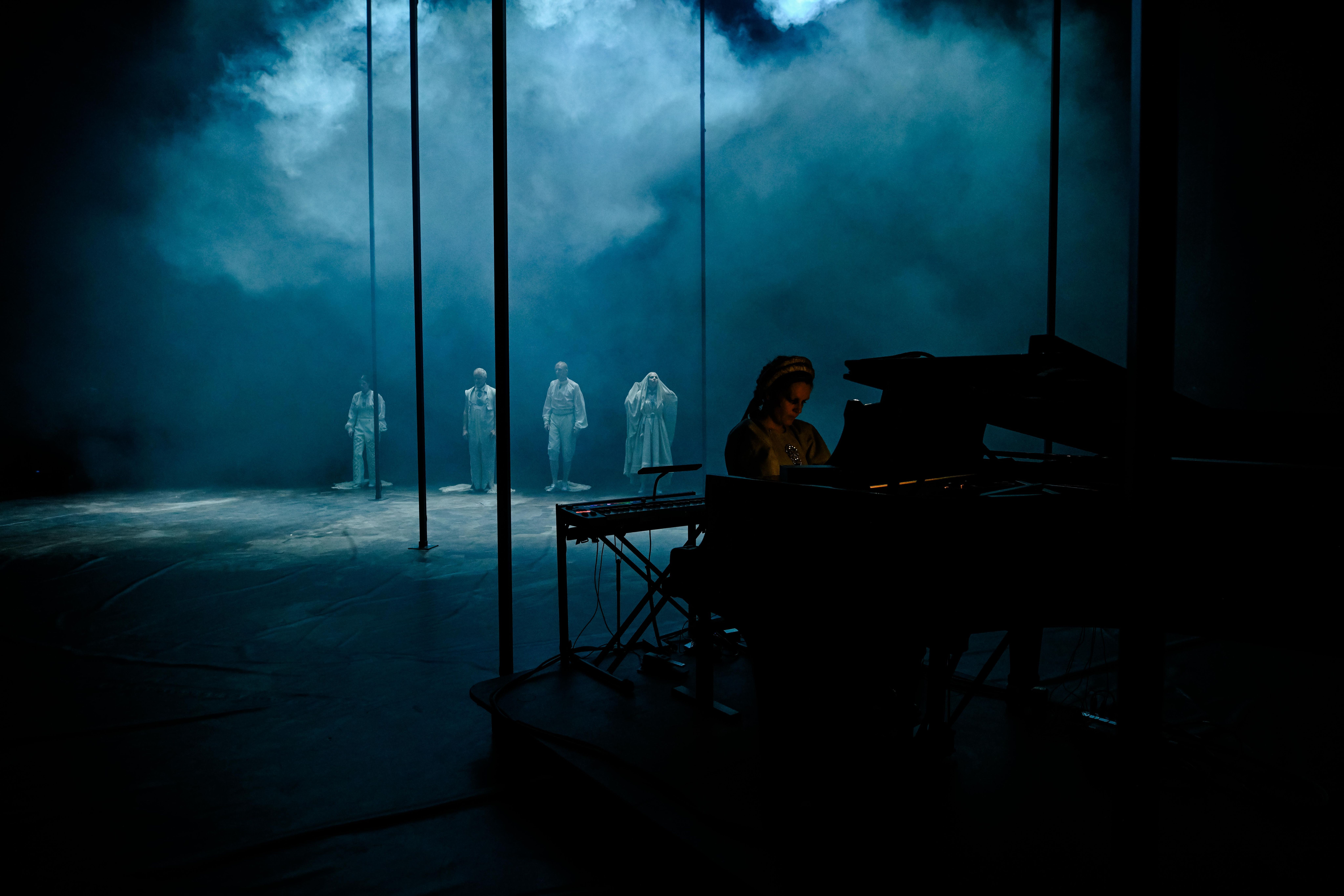 I forgrunnen en pianist ved flygel iført bunad. I bakgrunnen fire skuespillere i hvite kostymer på en mørk og røyklagt scene