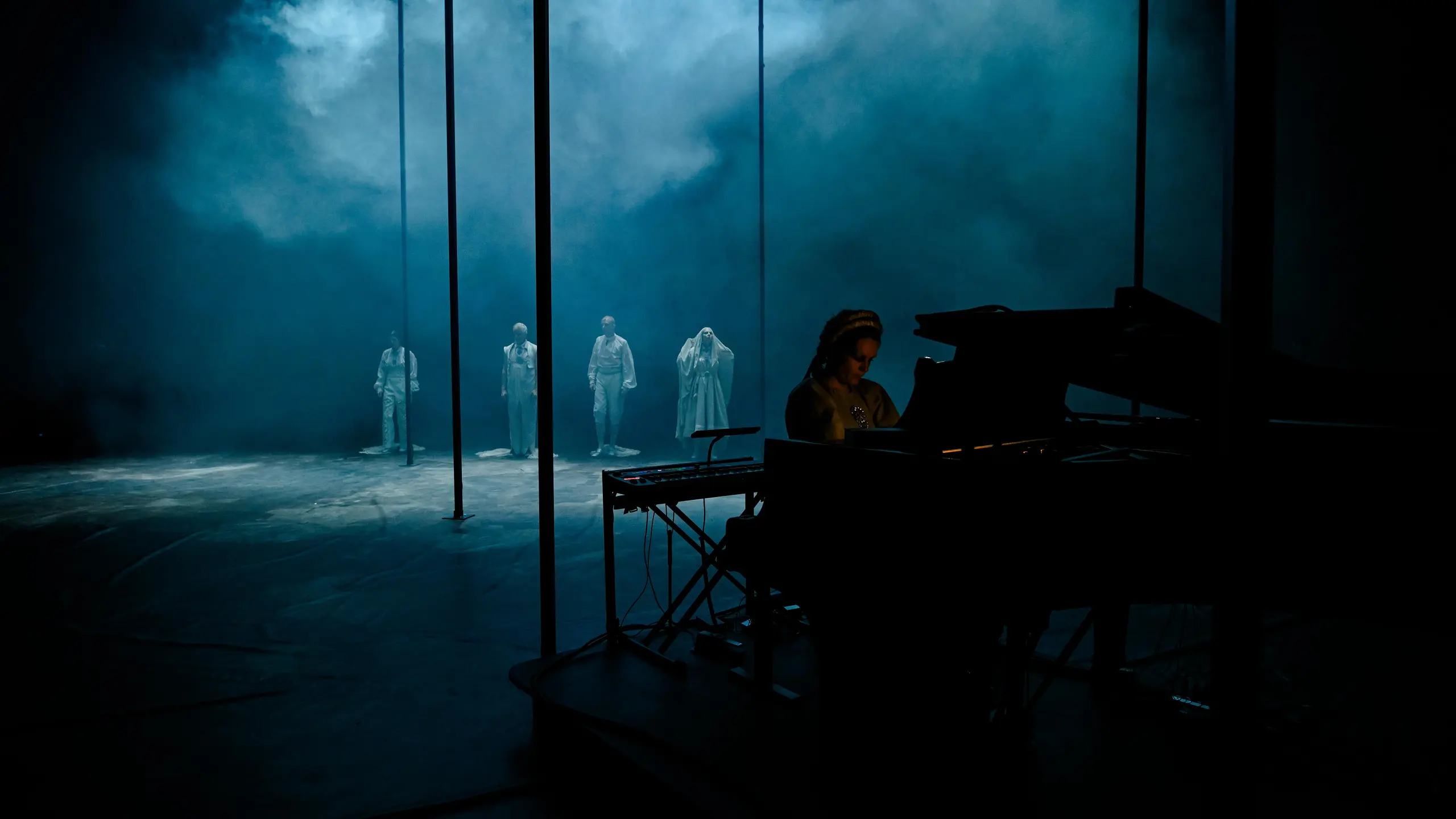 I forgrunnen en pianist ved flygel iført bunad. I bakgrunnen fire skuespillere i hvite kostymer på en mørk og røyklagt scene