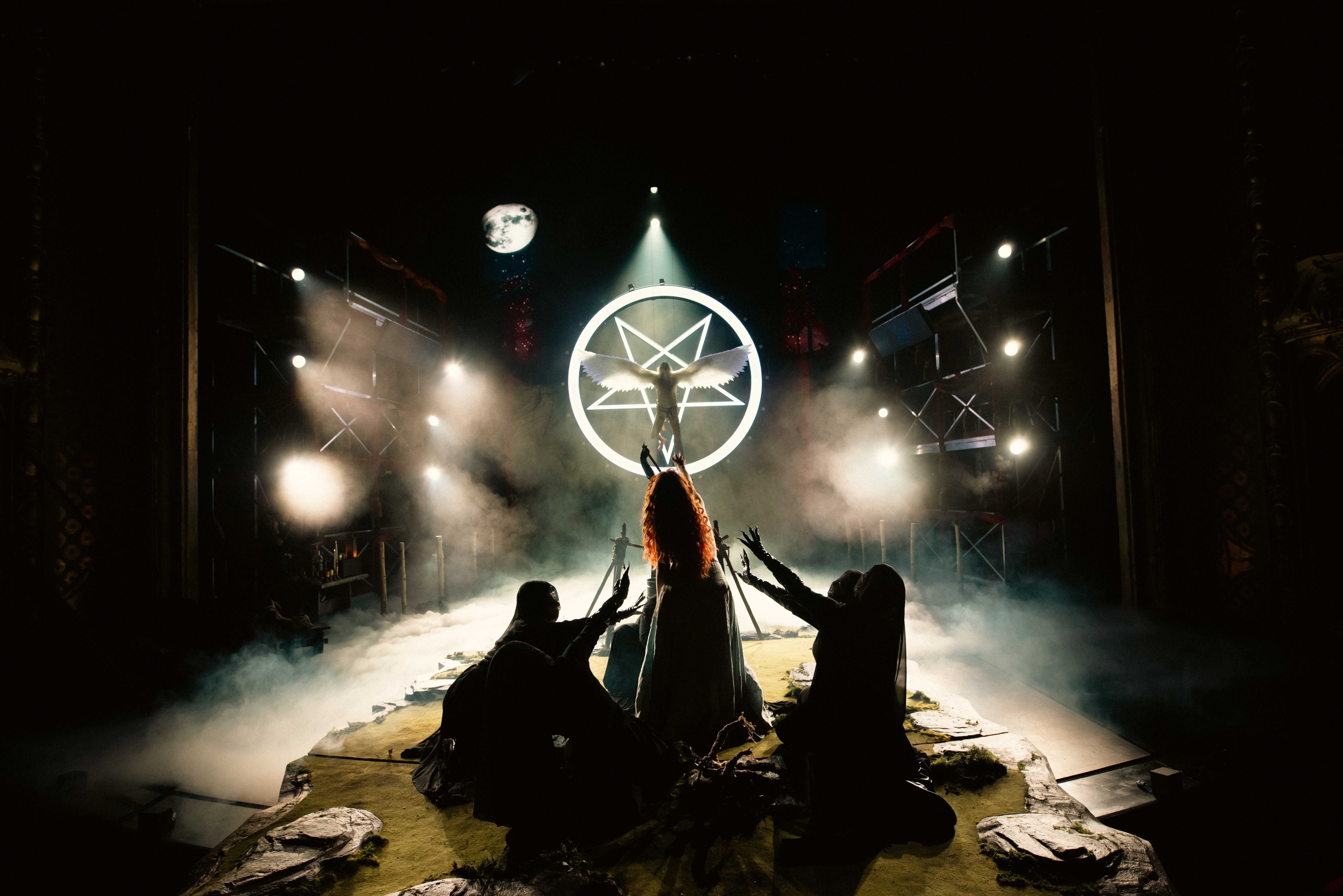 Baklyst scene med en bevinget figur foran en lysende pentagram-ring, omgitt av tre skikkelser som kneler med armene løftet.
