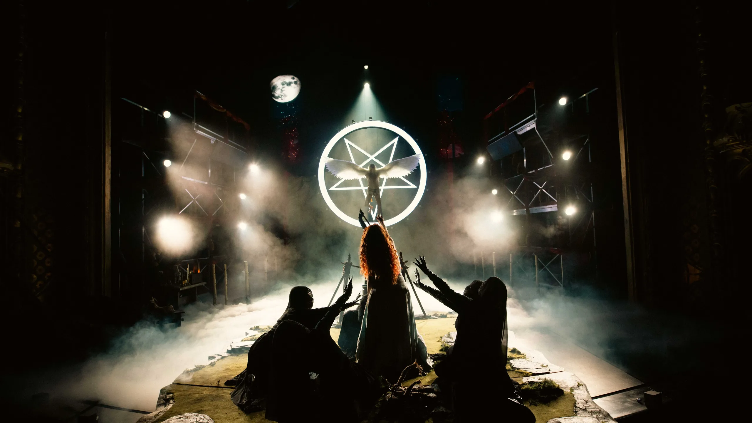 Baklyst scene med en bevinget figur foran en lysende pentagram-ring, omgitt av tre skikkelser som kneler med armene løftet.