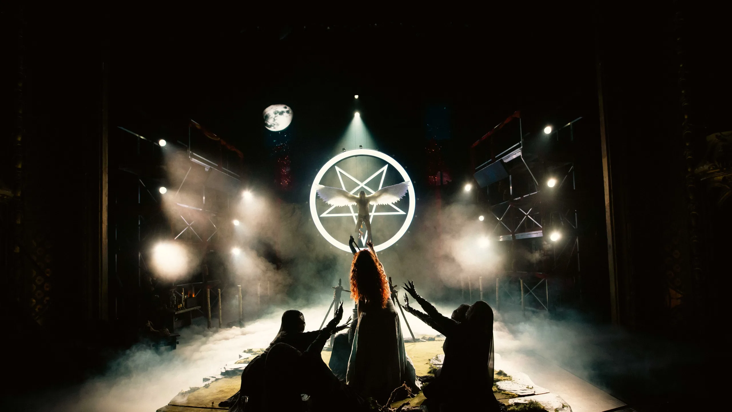 Baklyst scene med en bevinget figur foran en lysende pentagram-ring, omgitt av tre skikkelser som kneler med armene løftet.