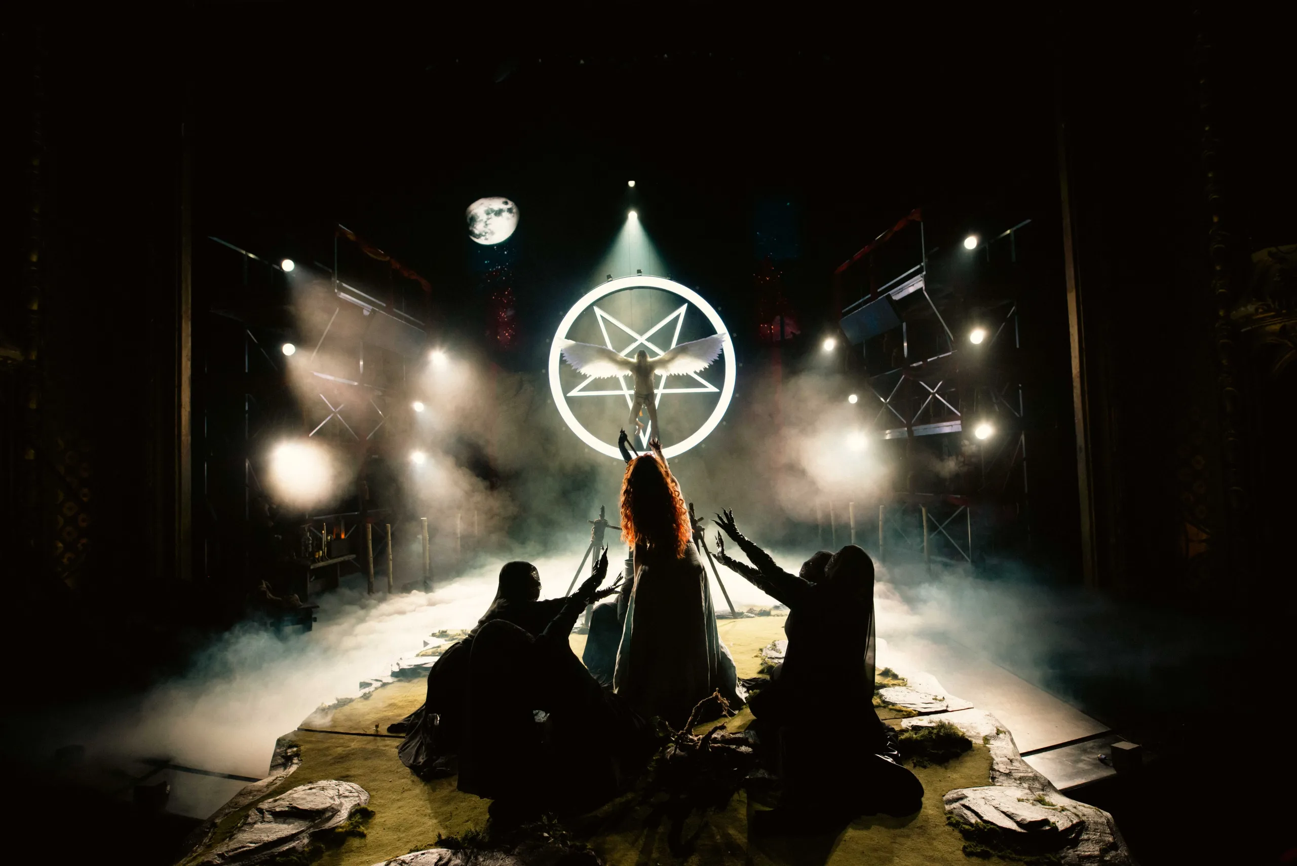 Baklyst scene med en bevinget figur foran en lysende pentagram-ring, omgitt av tre skikkelser som kneler med armene løftet.