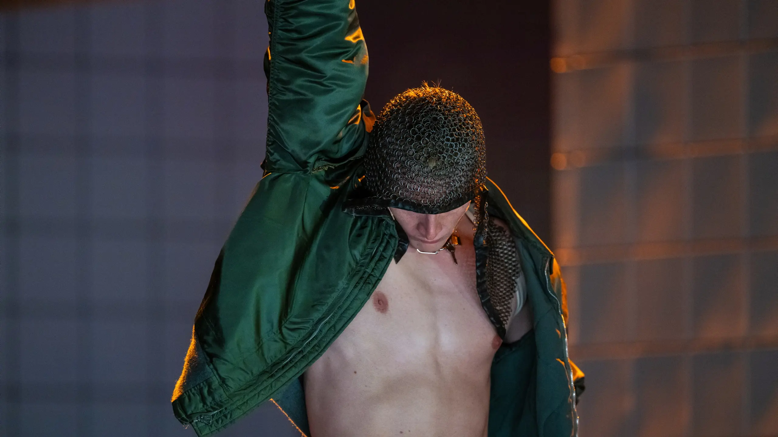 Skuespiller med bar overkropp og kjedekrave bøyer hodet ned, iført grønn jakke