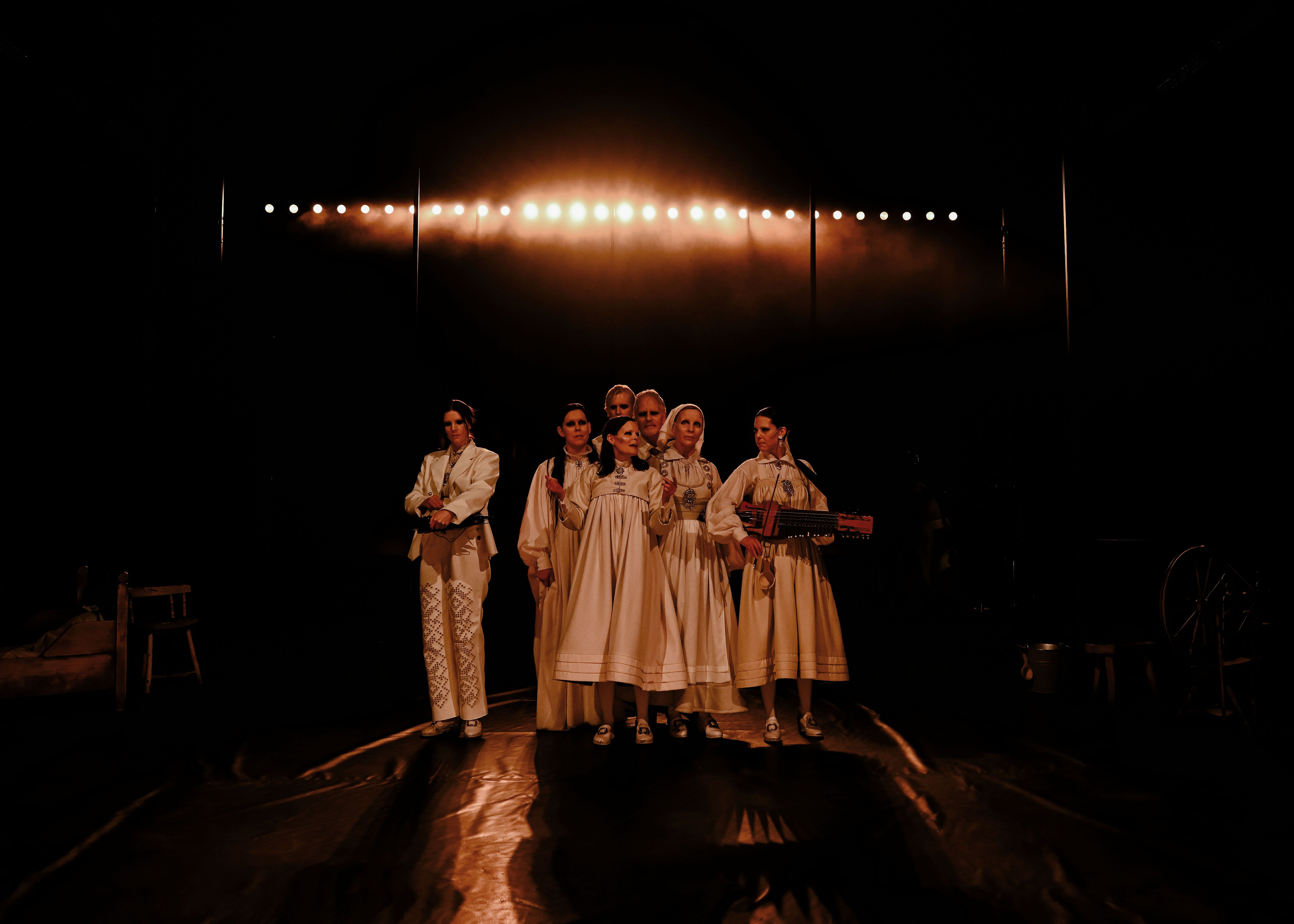 Syv aktører i bunadsinspirerte hvite kostymer med et par instrumenter på en mørk scene