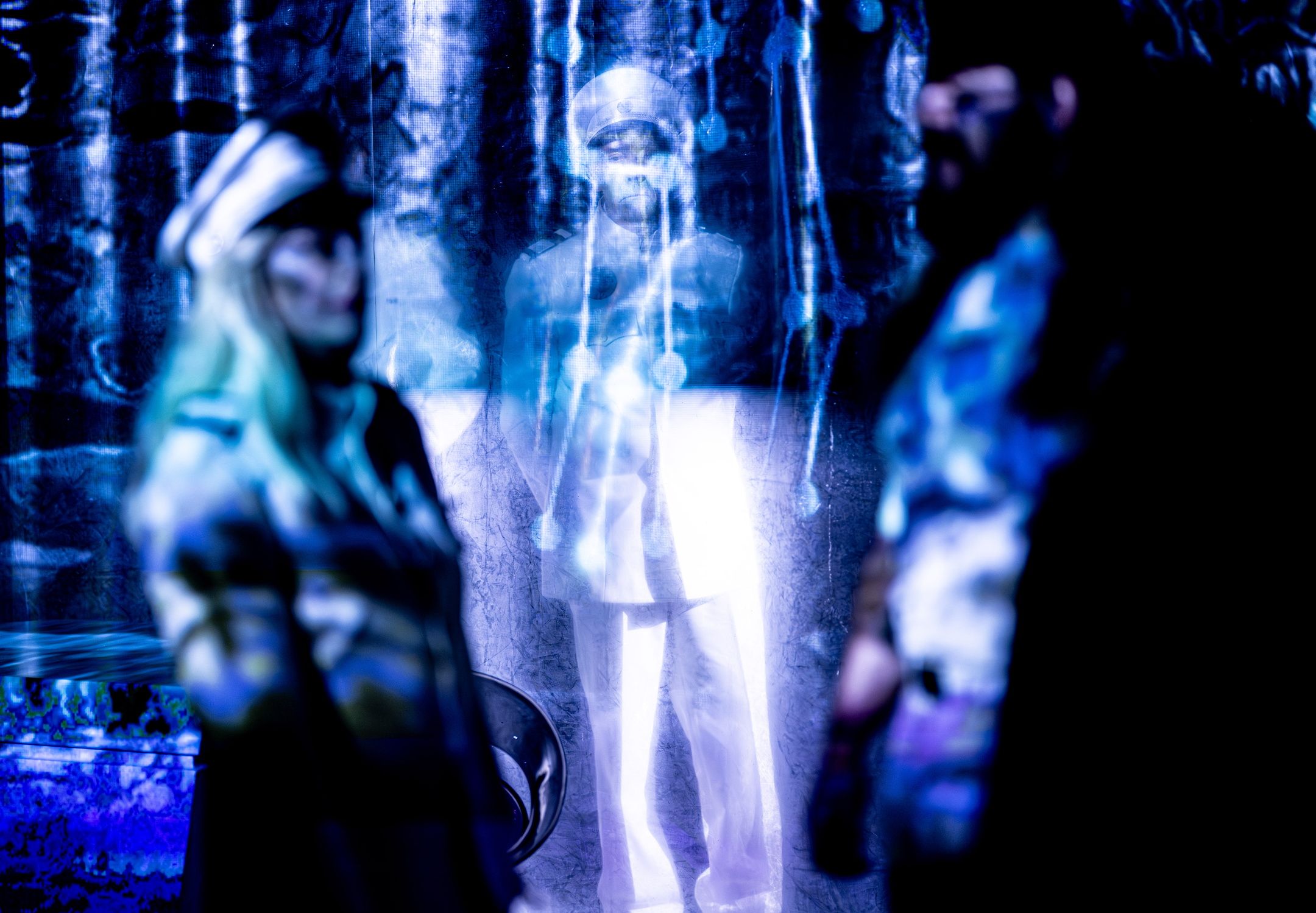 Lysende person i hvit uniform bak gjennomsiktig forheng, to uklare personer står i forgrunnen