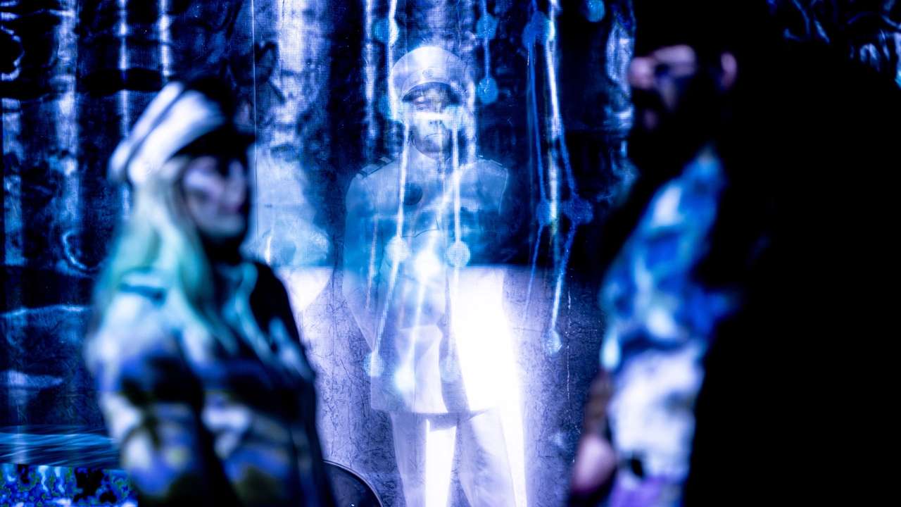 Lysende person i hvit uniform bak gjennomsiktig forheng, to uklare personer står i forgrunnen