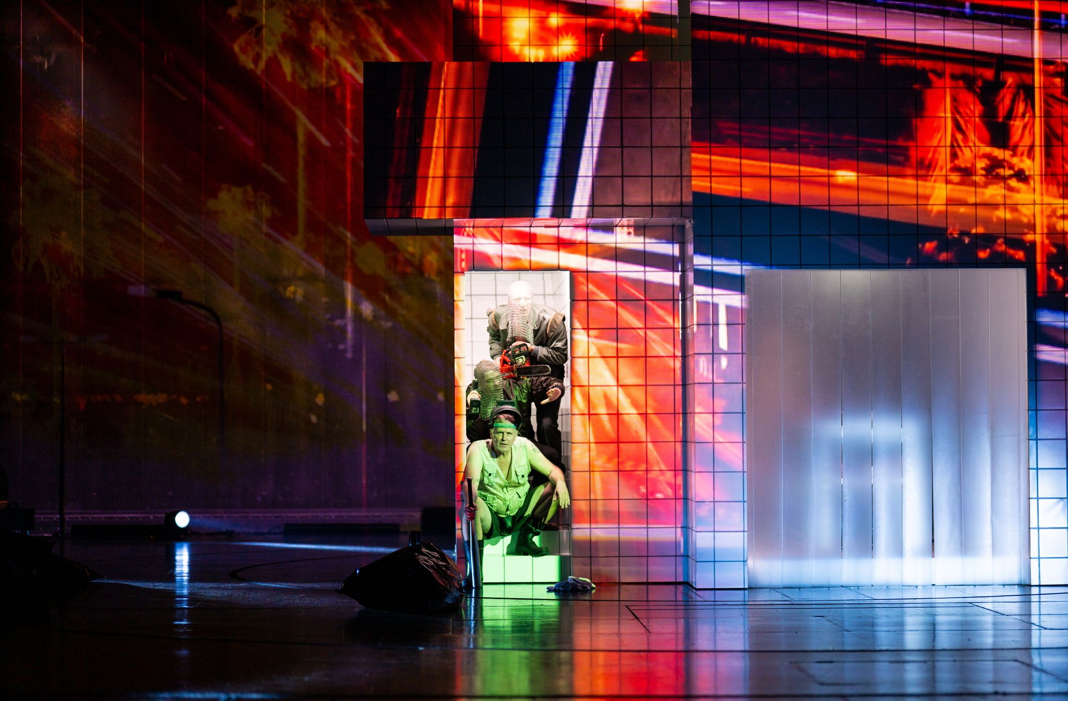 Tre personer i lagdelt sceneboks med digital lysprojeksjon og sterke farger, nederste person kledd i militærstil