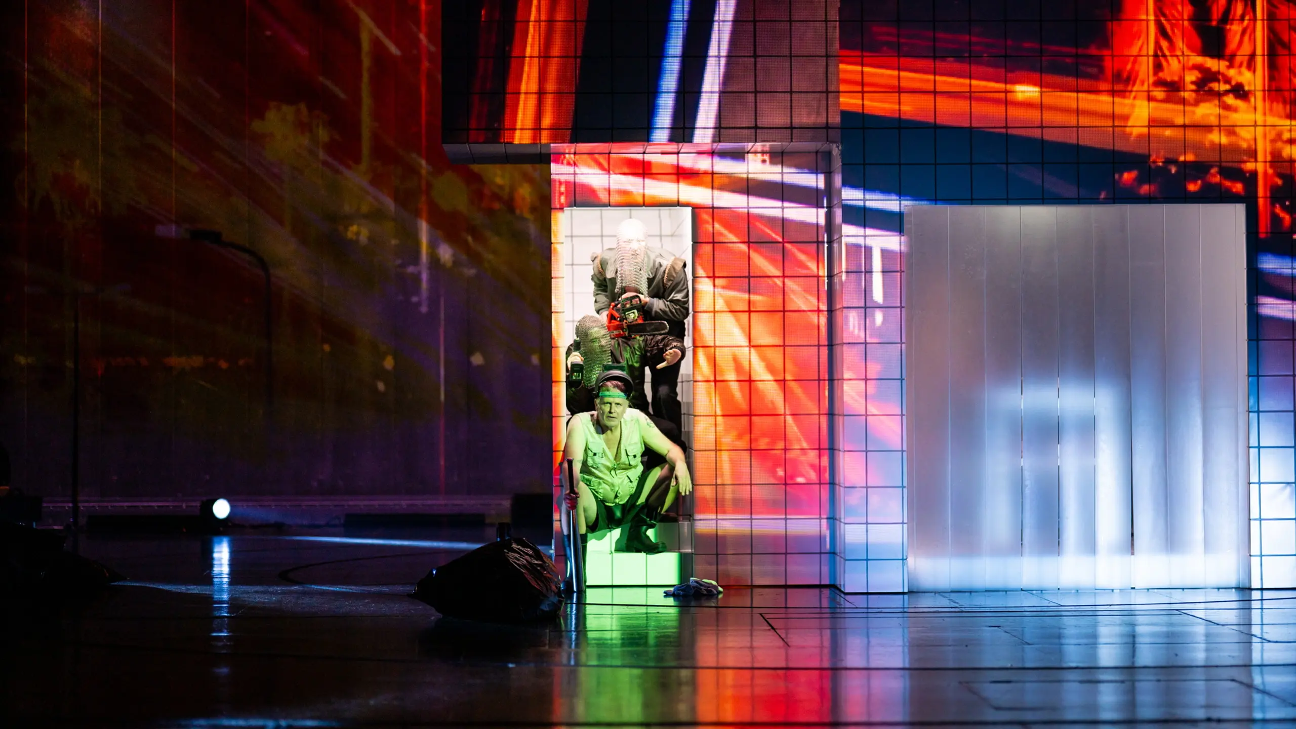 Tre personer i lagdelt sceneboks med digital lysprojeksjon og sterke farger, nederste person kledd i militærstil