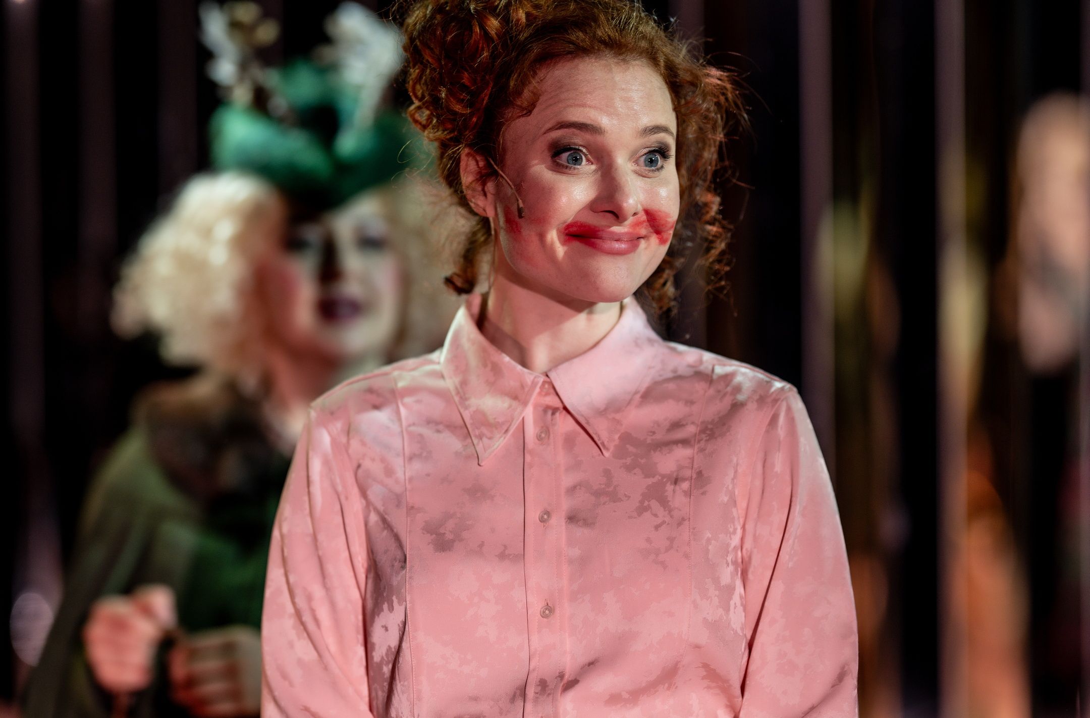 Kvinne i rosa skjorte med rød leppestift utover munnen smiler, mens annen figur i grønt er uskarp i bakgrunnen.