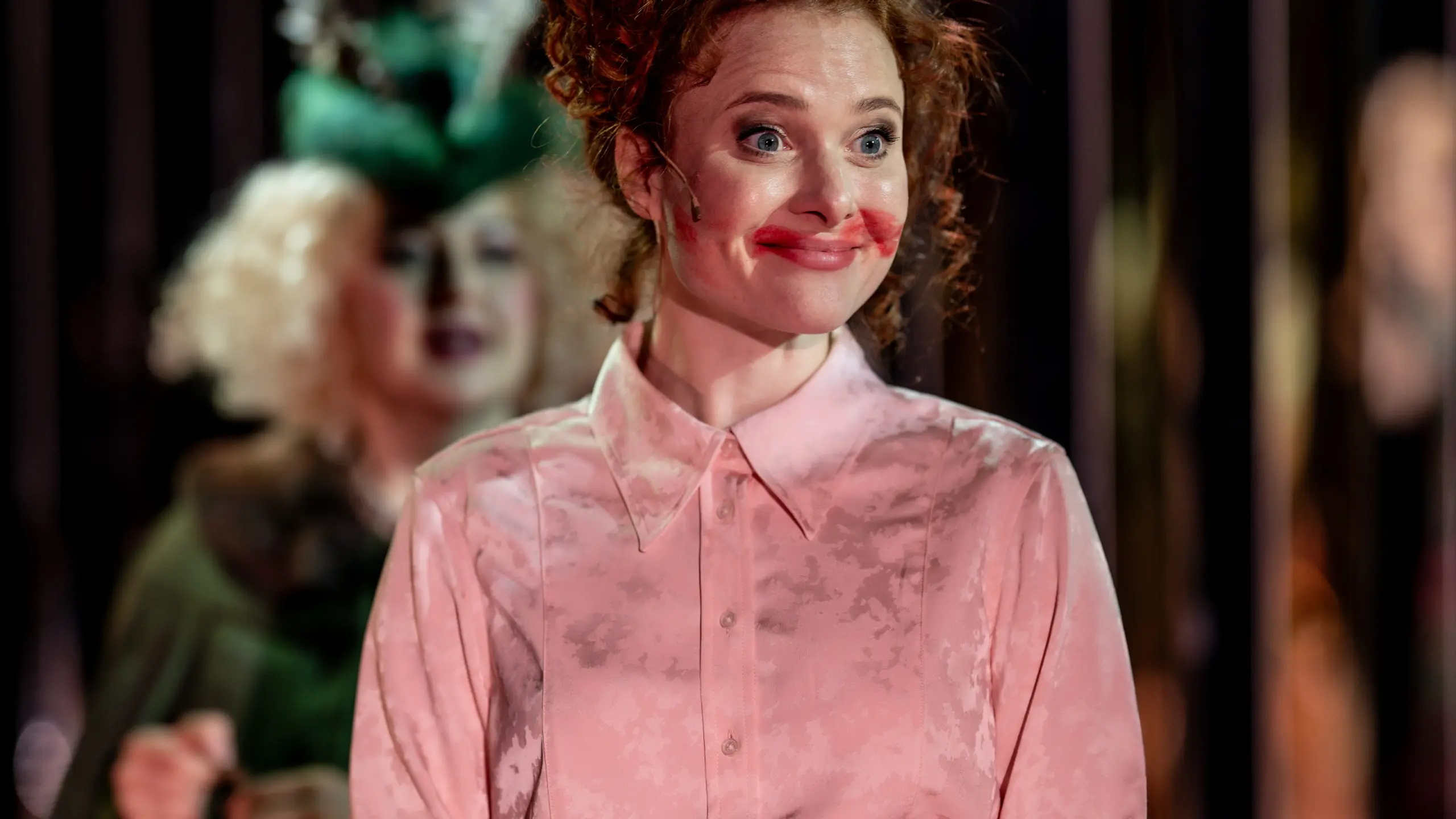 Kvinne i rosa skjorte med rød leppestift utover munnen smiler, mens annen figur i grønt er uskarp i bakgrunnen.