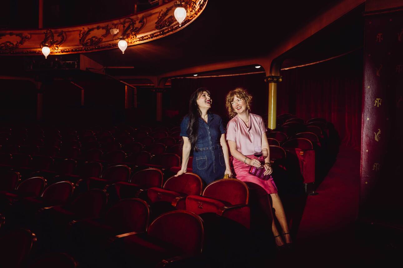 To kvinner sitter i en mørklagt teatersal med røde fløyelsseter og snakker og ler sammen. Den ene har på seg en mørkeblå kjole, den andre en rosa topp og skjørt, og holder et glass vin. De sitter avslappet ved siden av hverandre foran scenen, omgitt av teatrets klassiske, elegante interiør.