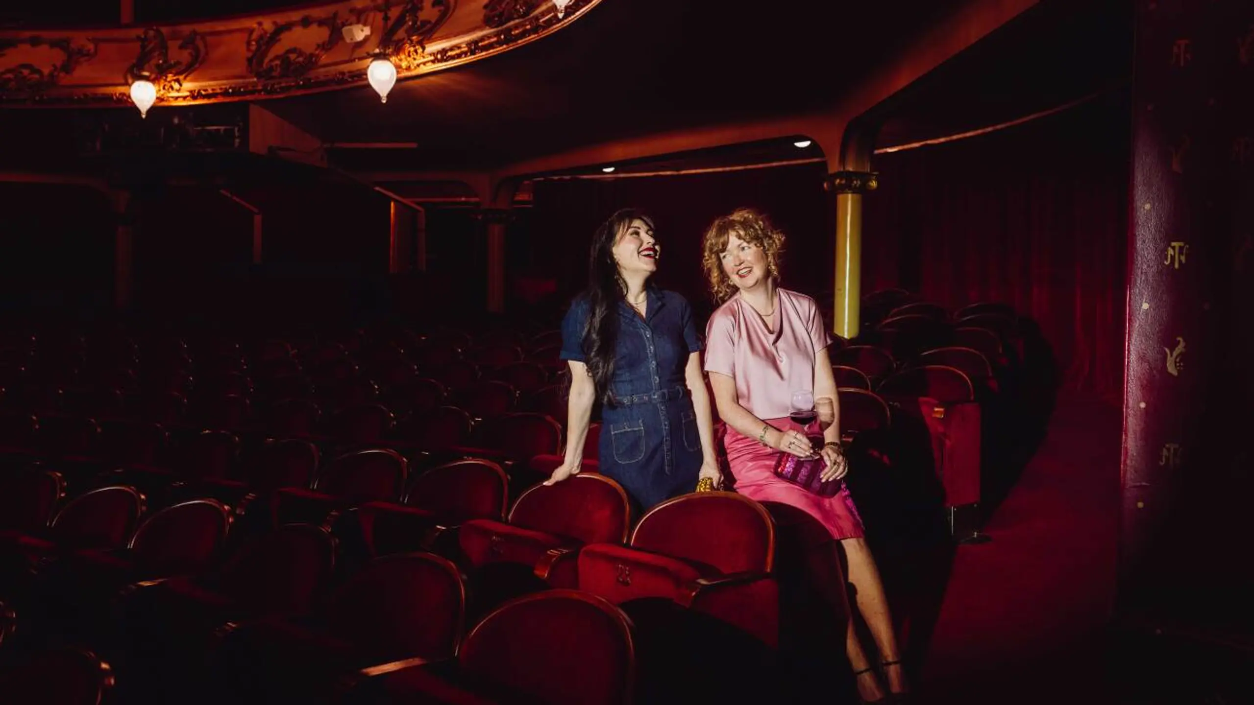 To kvinner sitter i en mørklagt teatersal med røde fløyelsseter og snakker og ler sammen. Den ene har på seg en mørkeblå kjole, den andre en rosa topp og skjørt, og holder et glass vin. De sitter avslappet ved siden av hverandre foran scenen, omgitt av teatrets klassiske, elegante interiør.