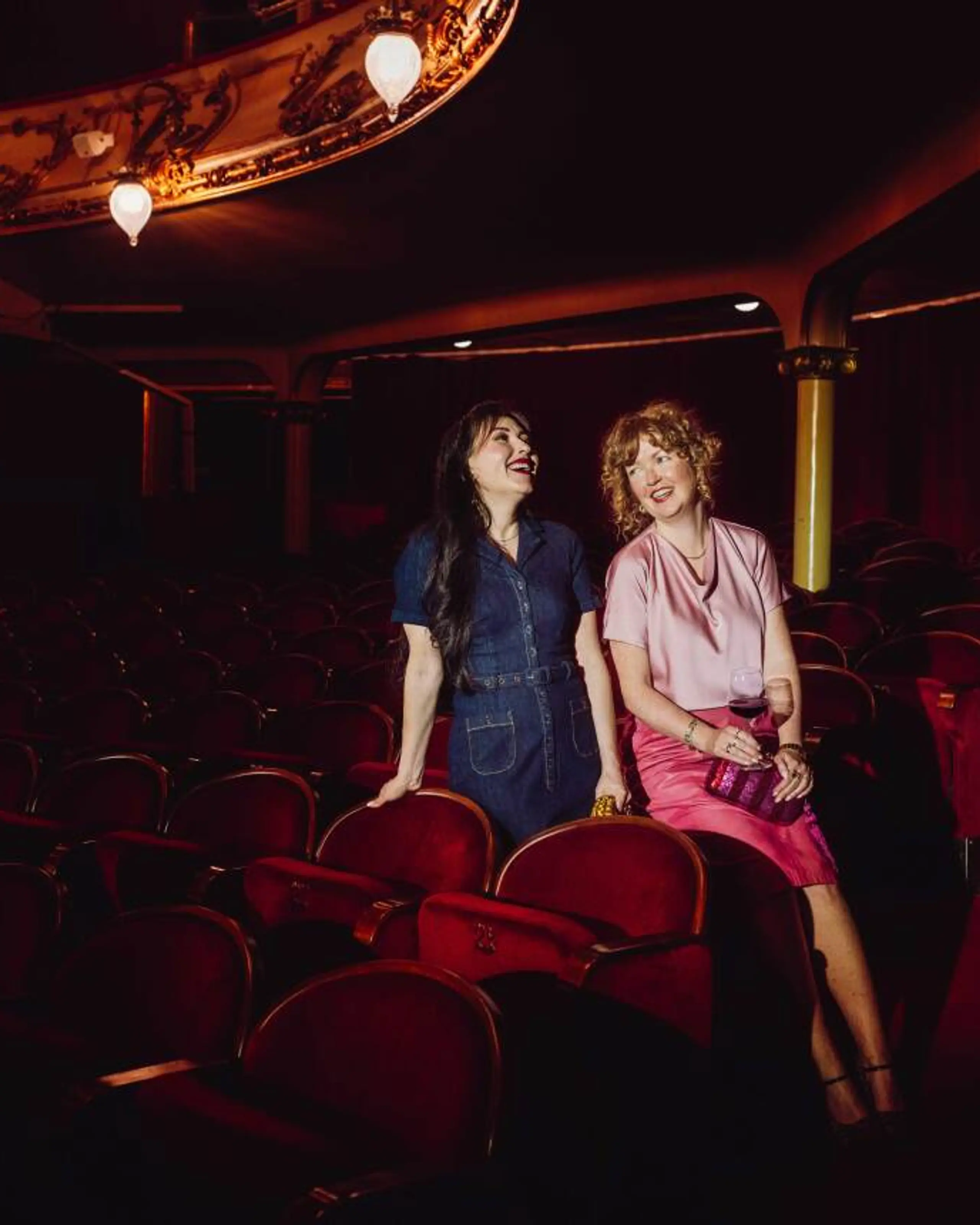 To kvinner sitter i en mørklagt teatersal med røde fløyelsseter og snakker og ler sammen. Den ene har på seg en mørkeblå kjole, den andre en rosa topp og skjørt, og holder et glass vin. De sitter avslappet ved siden av hverandre foran scenen, omgitt av teatrets klassiske, elegante interiør.
