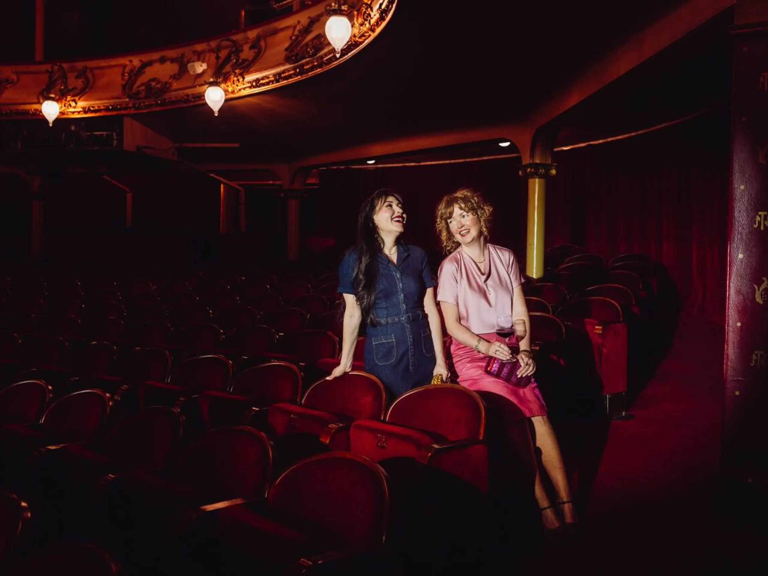 To kvinner sitter i en mørklagt teatersal med røde fløyelsseter og snakker og ler sammen. Den ene har på seg en mørkeblå kjole, den andre en rosa topp og skjørt, og holder et glass vin. De sitter avslappet ved siden av hverandre foran scenen, omgitt av teatrets klassiske, elegante interiør.