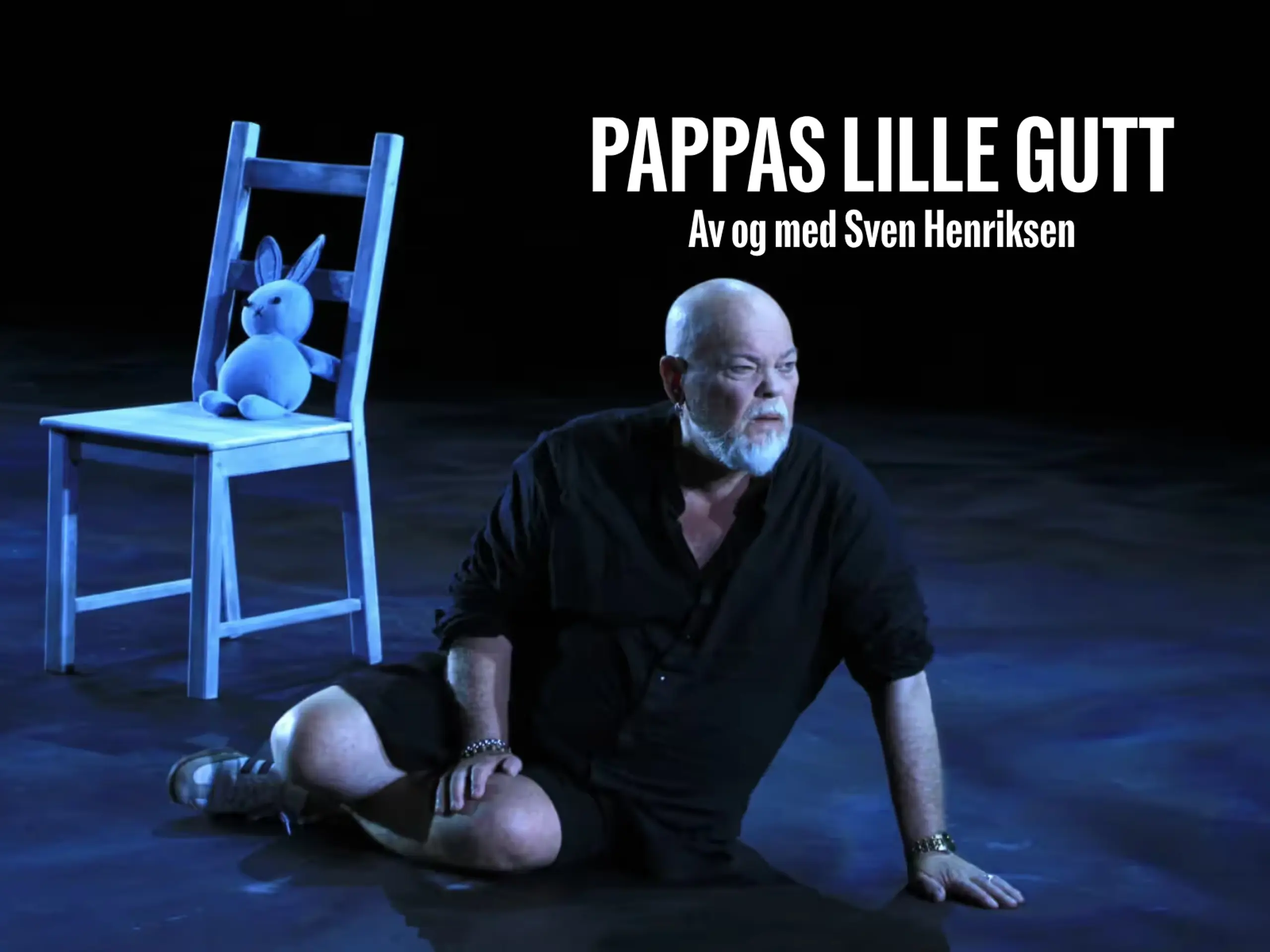 Mann sitter på gulvet på en mørk scene. Ved siden av ham står en hvit pinnestol med en bamse på. Med tekst Pappas lille gutt av og med Sven Henriksen