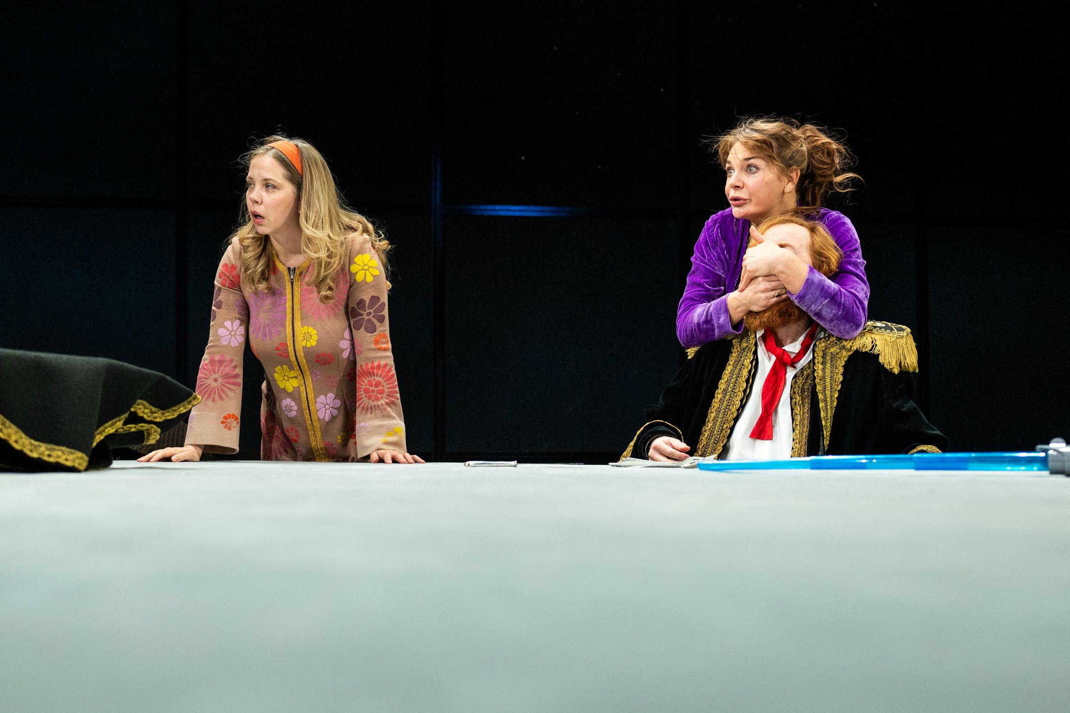 Kvinne i lilla holder hånden over munnen på en mann i uniform, mens en annen kvinne ser forskrekket på.