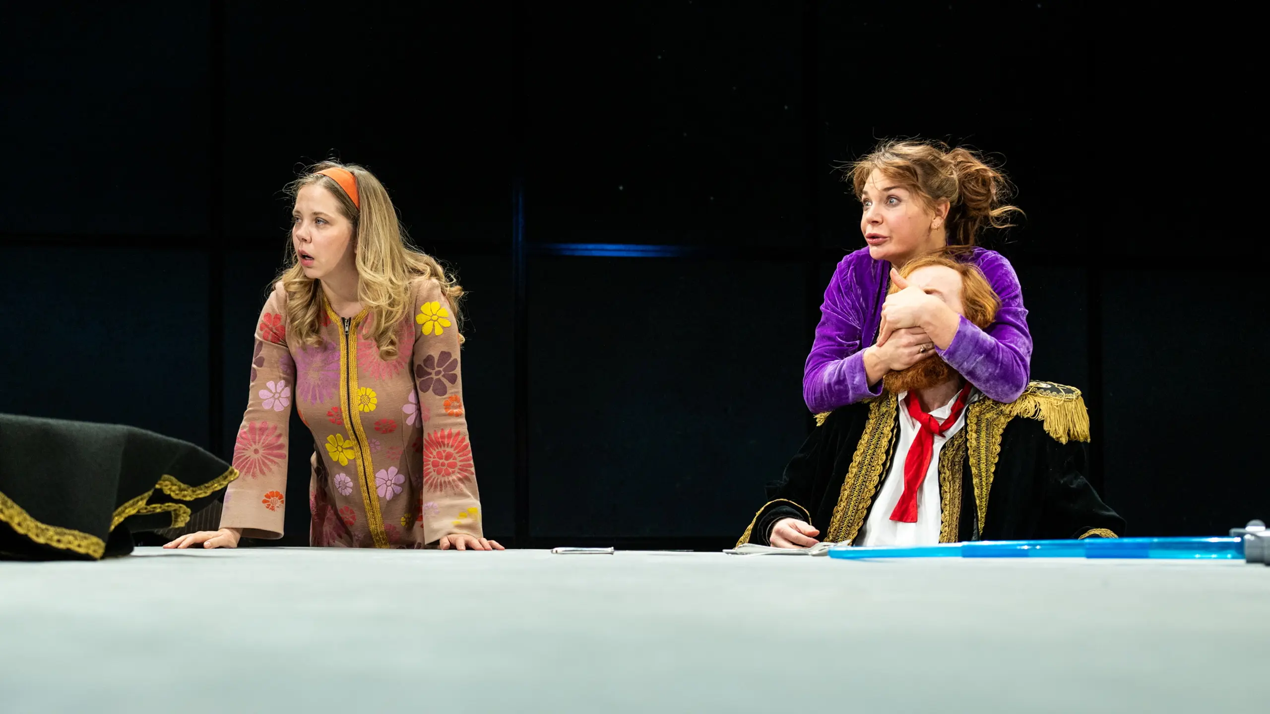 Kvinne i lilla holder hånden over munnen på en mann i uniform, mens en annen kvinne ser forskrekket på.