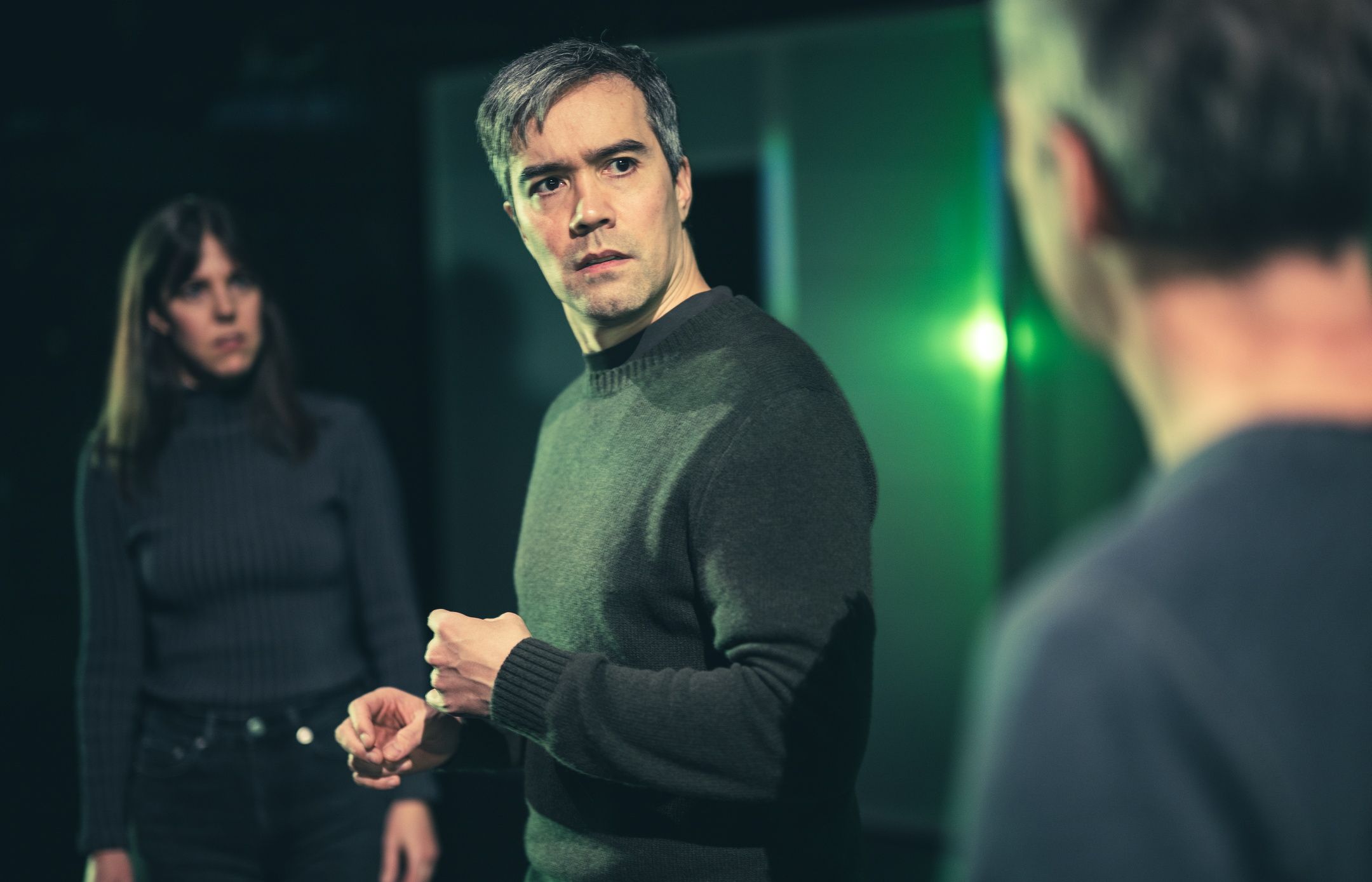 En mann ser bekymret på en annen mann i forgrunnen, mens en kvinne i bakgrunnen ser på dem med alvorlig blikk.