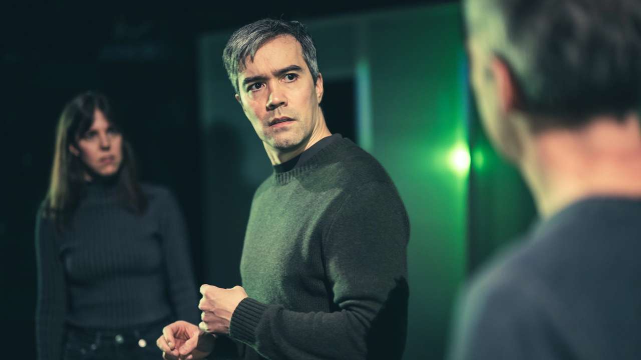 En mann ser bekymret på en annen mann i forgrunnen, mens en kvinne i bakgrunnen ser på dem med alvorlig blikk.