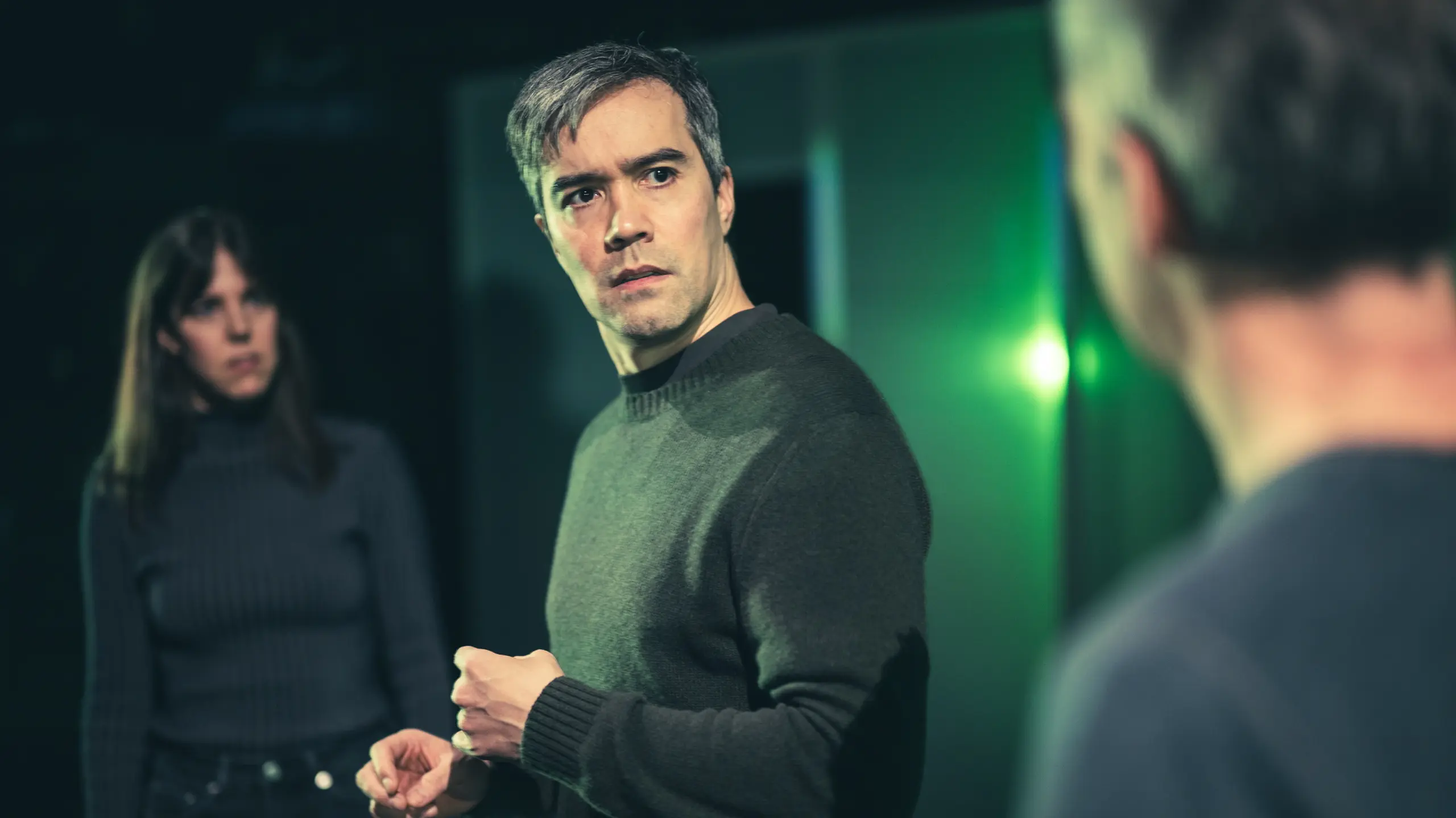 En mann ser bekymret på en annen mann i forgrunnen, mens en kvinne i bakgrunnen ser på dem med alvorlig blikk.