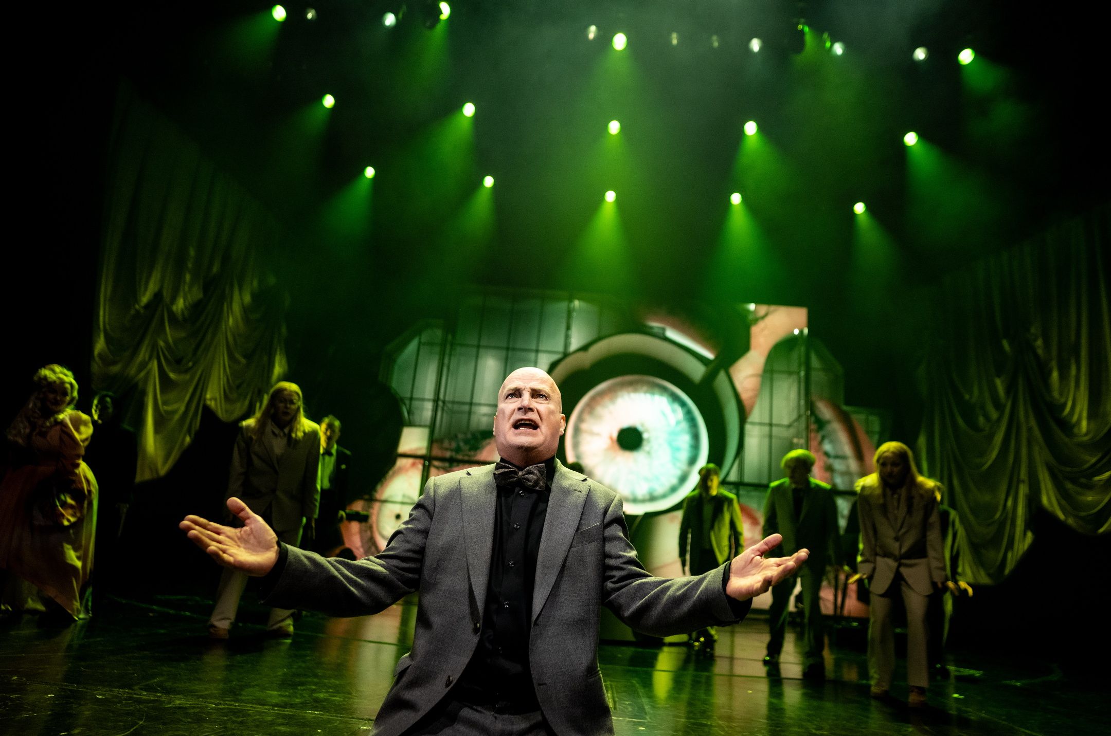 Skallet person i grå dress gestikulerer foran gruppe aktører under grønne scenelys og øye-projeksjon bak.