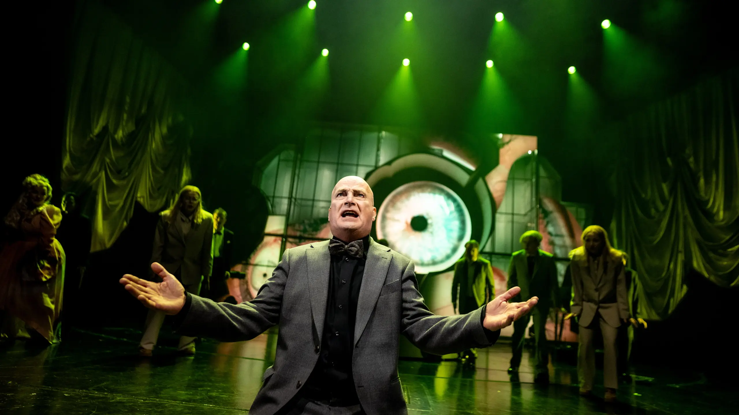 Skallet person i grå dress gestikulerer foran gruppe aktører under grønne scenelys og øye-projeksjon bak.