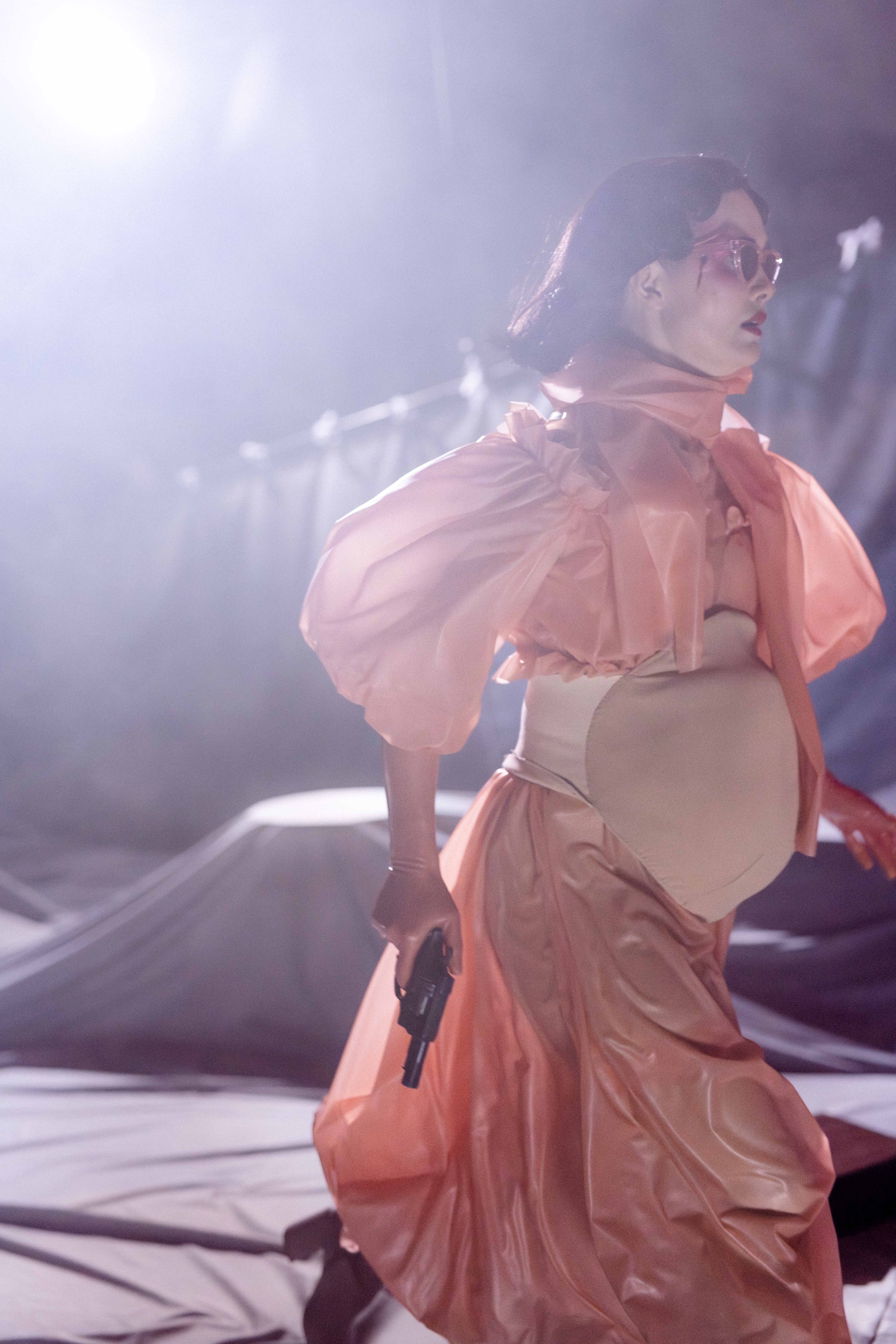 Person i ferskenfarget kjole og solbriller går over scenen mens hun holder en pistol i hånden